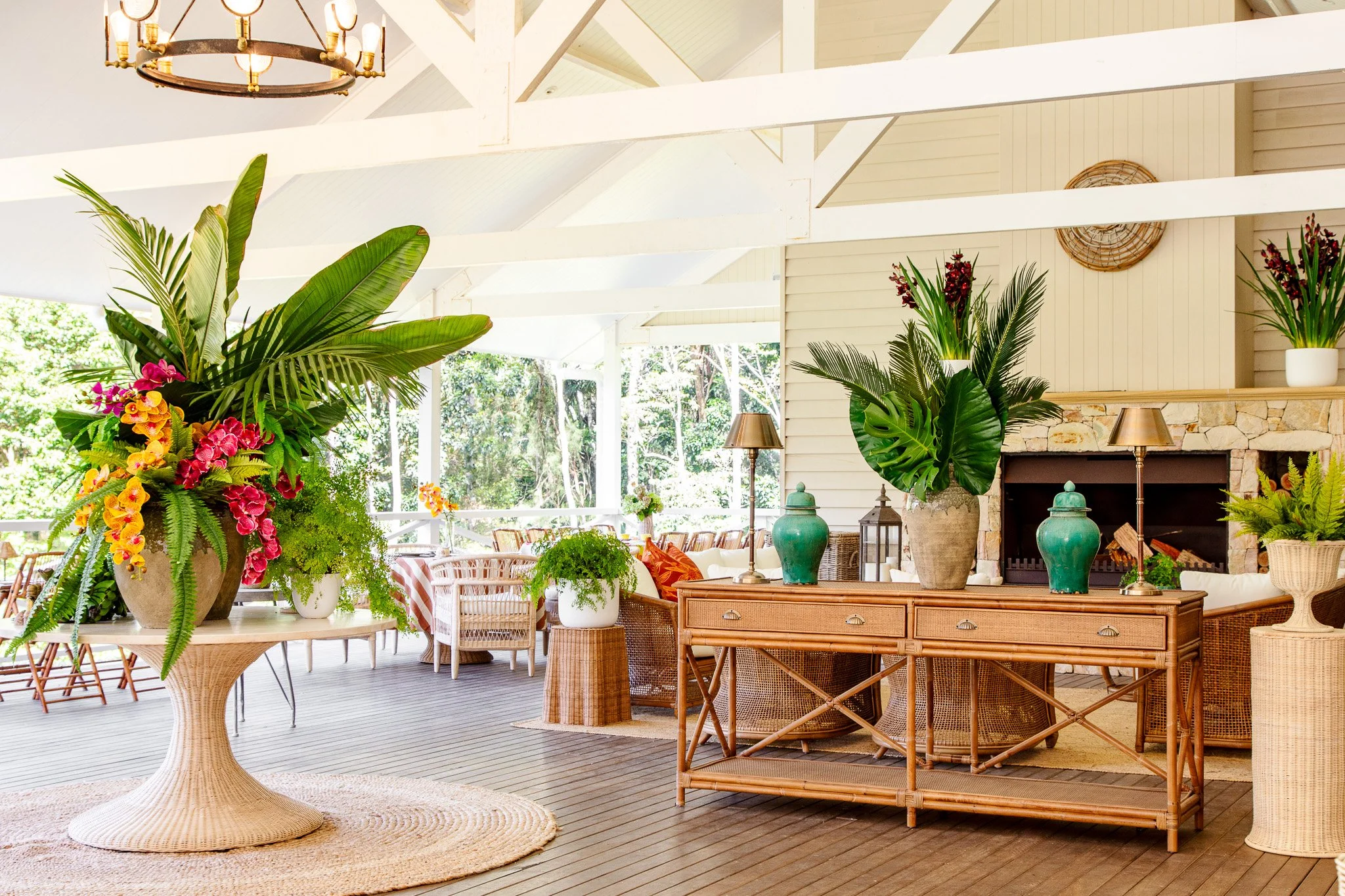 Bright indoor lounge with large floral arrangements, wicker furniture, fireplace, potted plants, and large windows showing greenery outside.