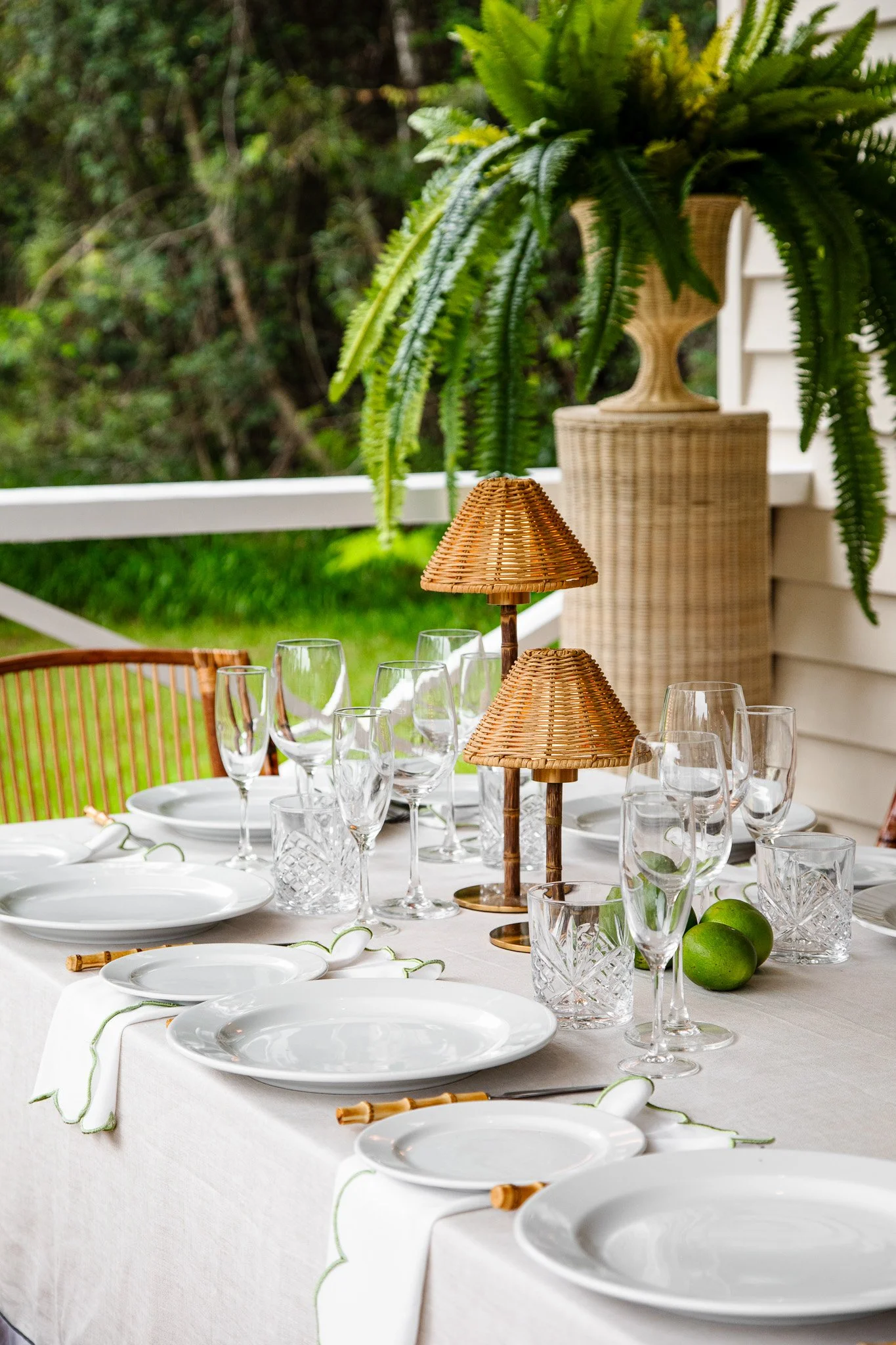 A beautifully set outdoor dining table with white plates, crystal glasses, wicker lamps, and fresh limes, surrounded by lush greenery.