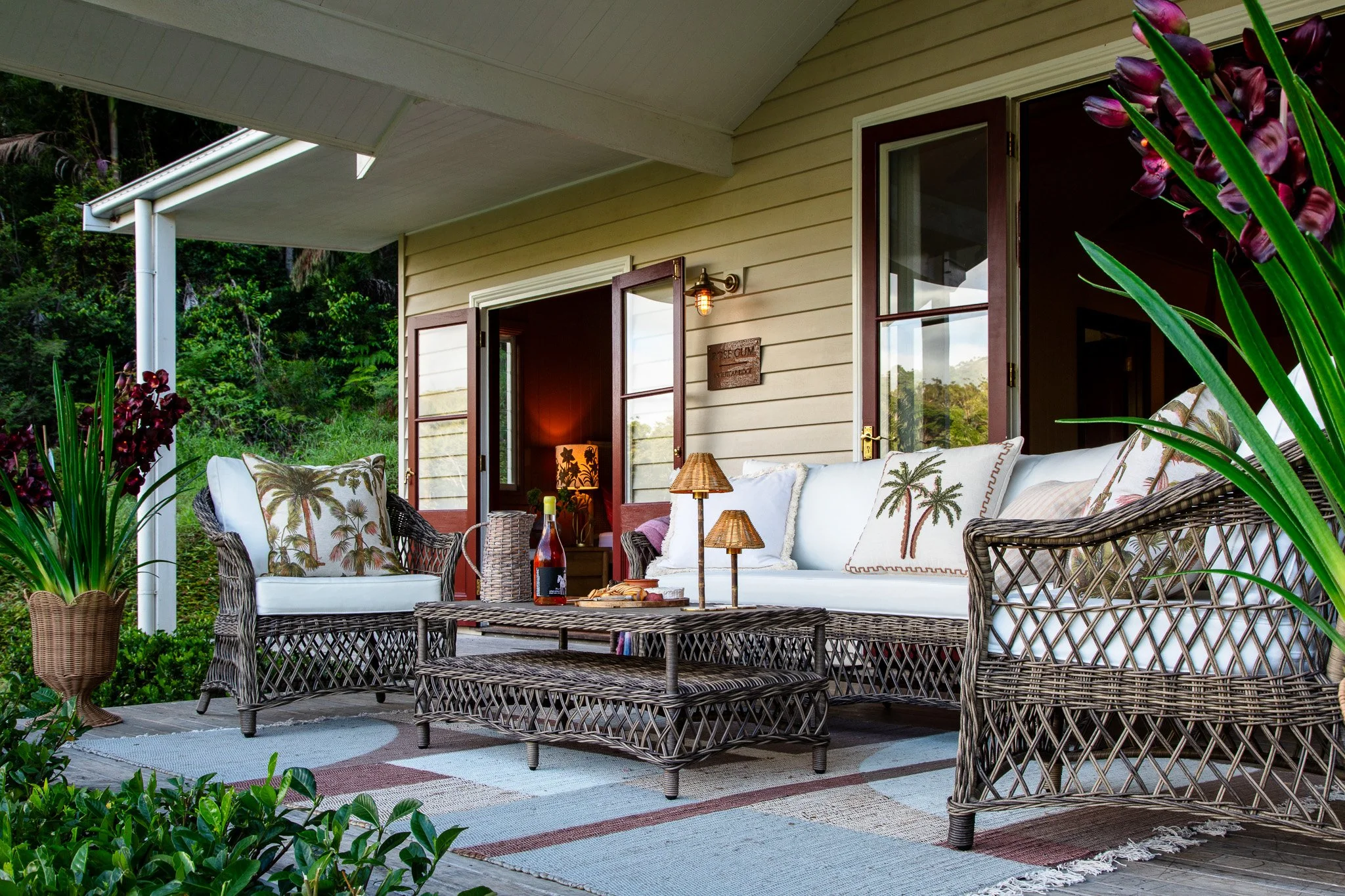 Outdoor patio with wicker furniture, white cushions, decorative pillows with palm tree prints, a woven table with a bottle of wine, lamps, and lush greenery surrounding a house with beige siding and open doors.