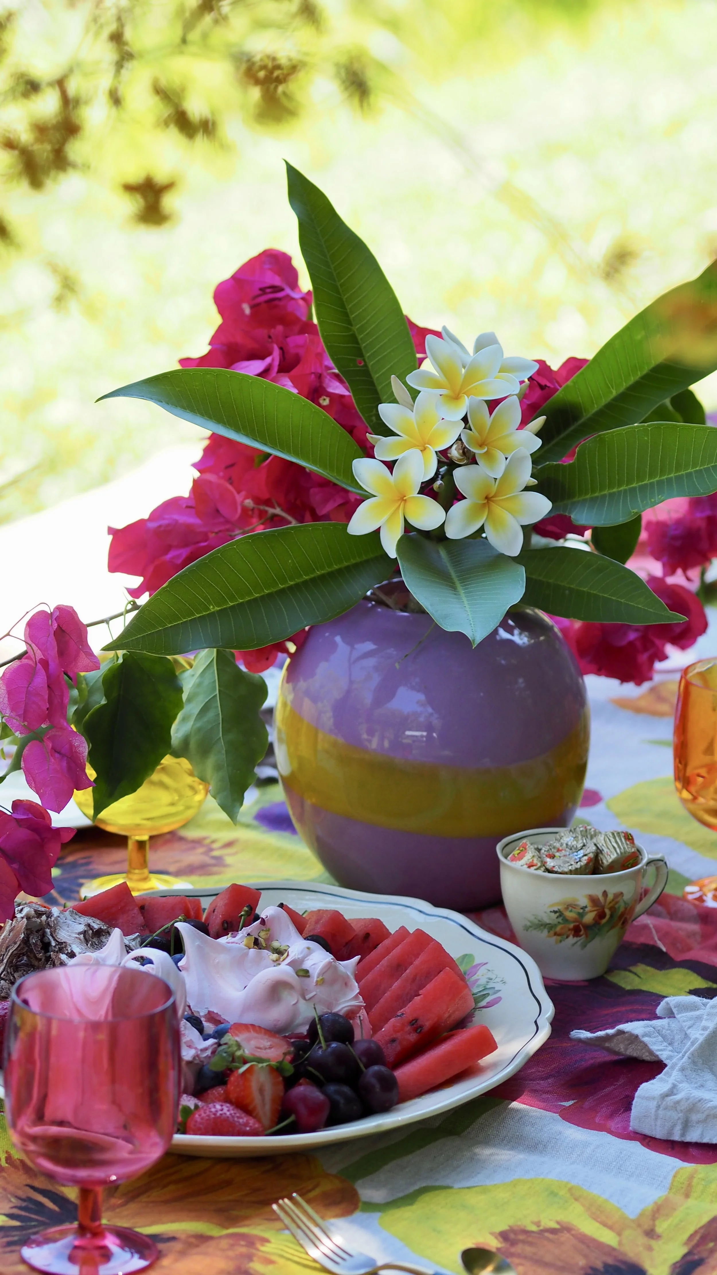 Colorful table setting with a large purple vase holding pink and white flowers and green leaves, surrounded by pink, yellow, and orange glasses, a plate of watermelon, strawberries, cherries, and a dollop of whipped cream, and a small cup with wrapped candies in a sunny outdoor setting.