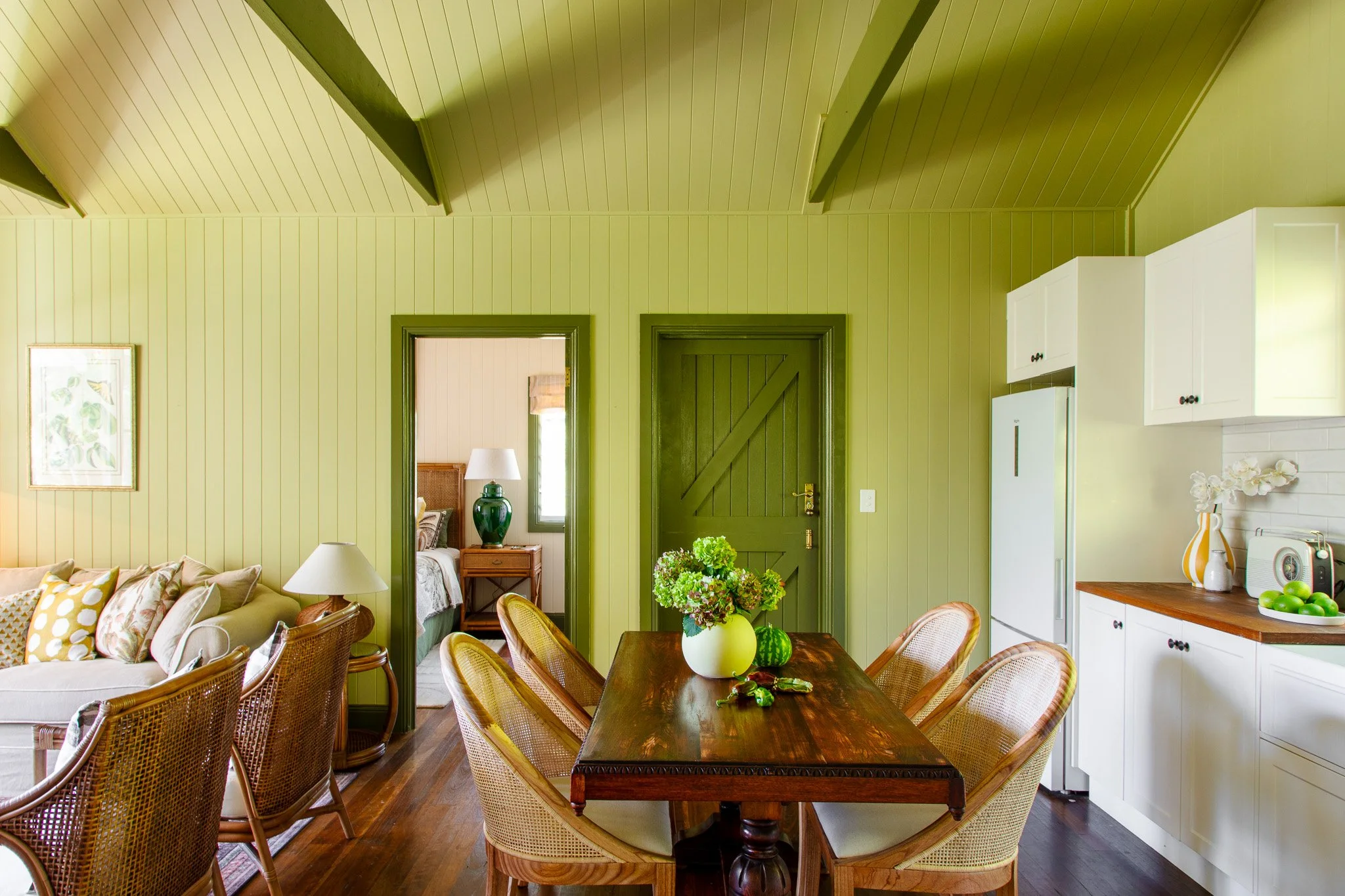 Interior of a dining room with green paneled walls and ceiling, wooden dining table with a flower vase, rattan chairs around the table, adjacent living room with beige sofa and artwork, and a view into a bedroom with a bed and nightstand through an o