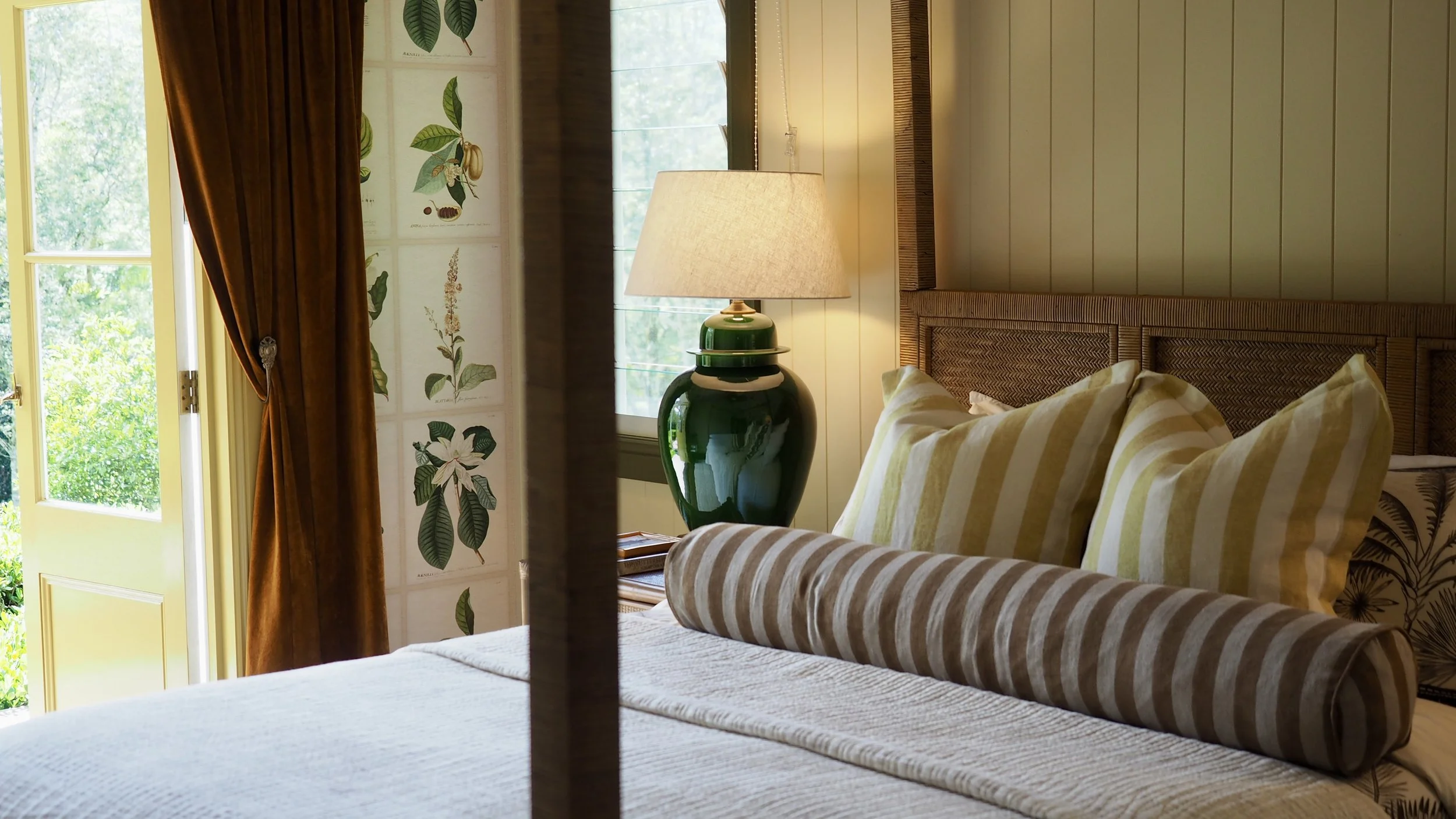 A cozy bedroom with a bed, striped pillows, a green lamp on a bedside table, a window with natural light, and a floral wall decoration.