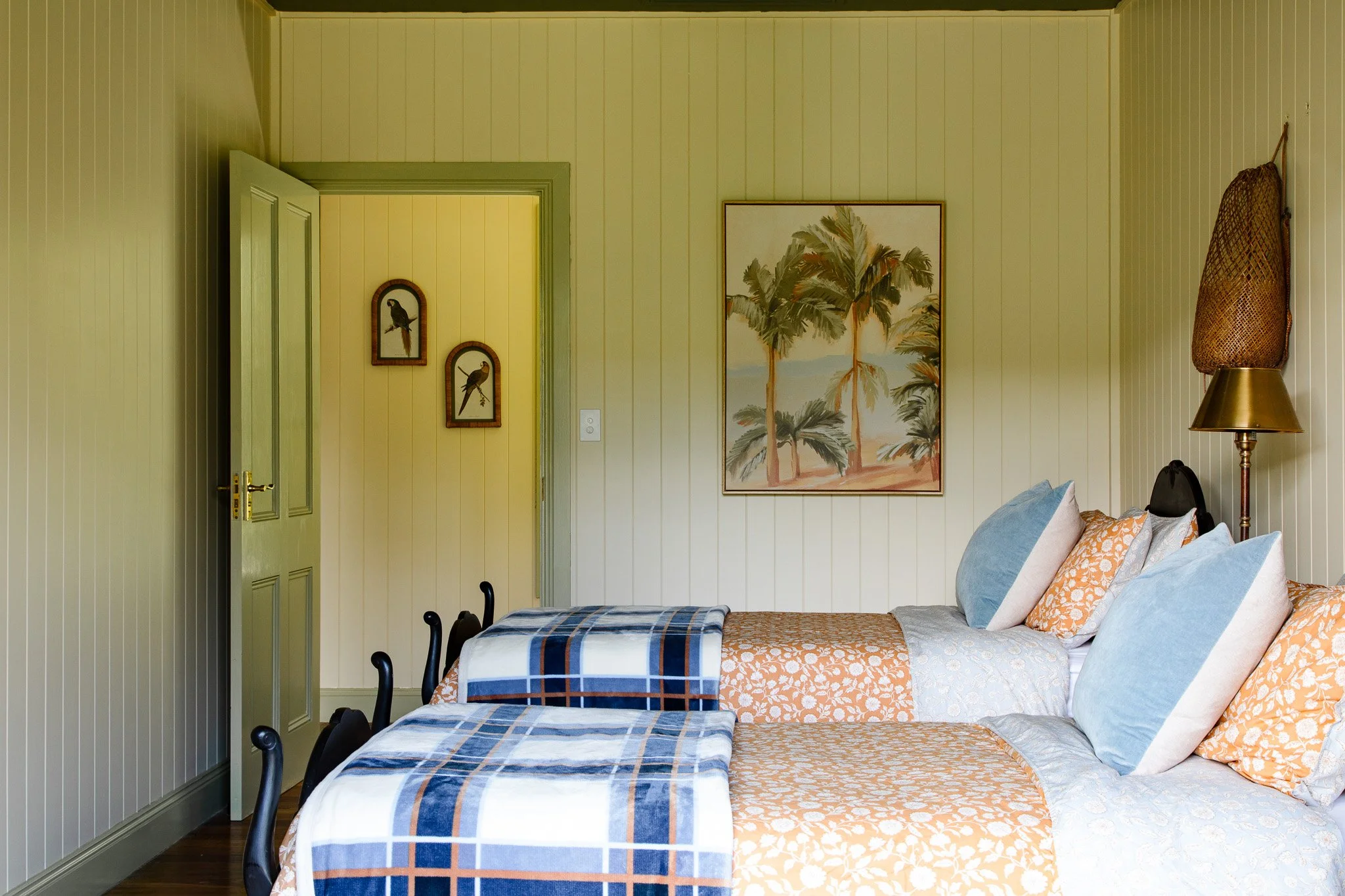 Two beds with blue and orange bedding, white and orange pillows, in a room with beige paneled walls. There's a painting of palm trees, a lamp with a woven cover, and a door with bird-themed wall art.