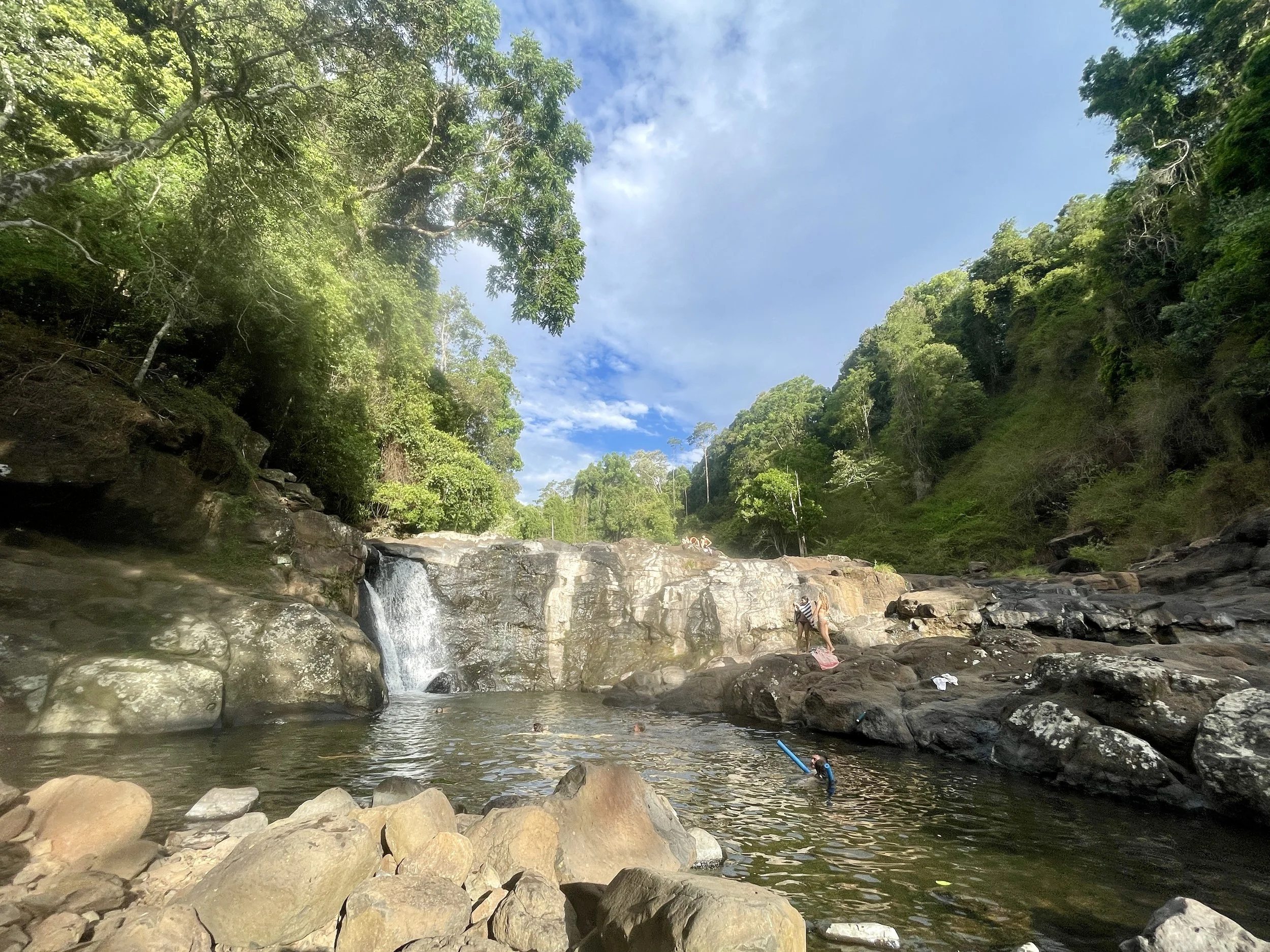 A scenic river with small waterfall surrounded by lush green trees on both sides, under a partly cloudy sky, with people relaxing and swimming near the rocks.