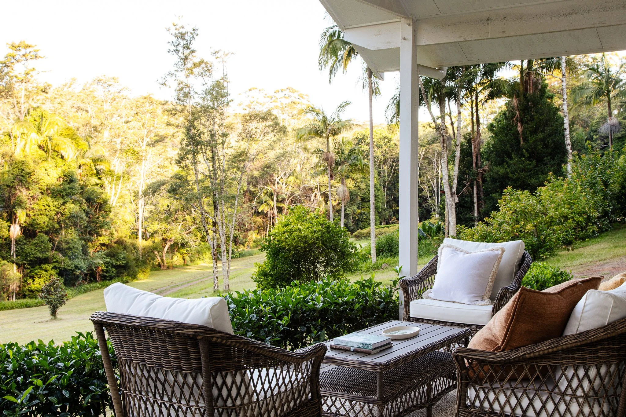 Outdoor patio with wicker furniture including chairs and a table, surrounded by lush greenery and trees in the background.