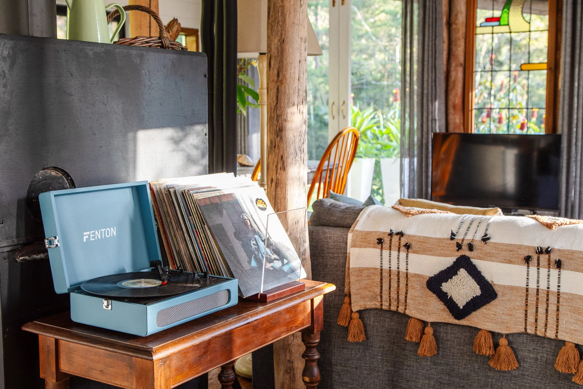 A vintage turntable on a wooden table, vinyl records, a gray armchair with a beige and black woven throw blanket, a flat-screen TV, and a bright sunlit room with large windows and green plants outside.