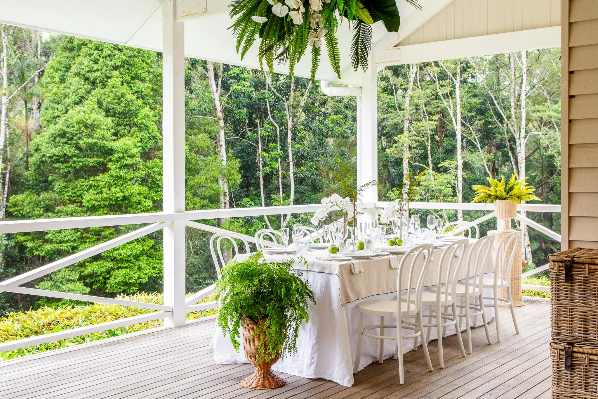 Outdoor dining table set on a porch overlooking a lush green forest with white chairs, tablecloth, floral centerpiece, and greenery decorations.