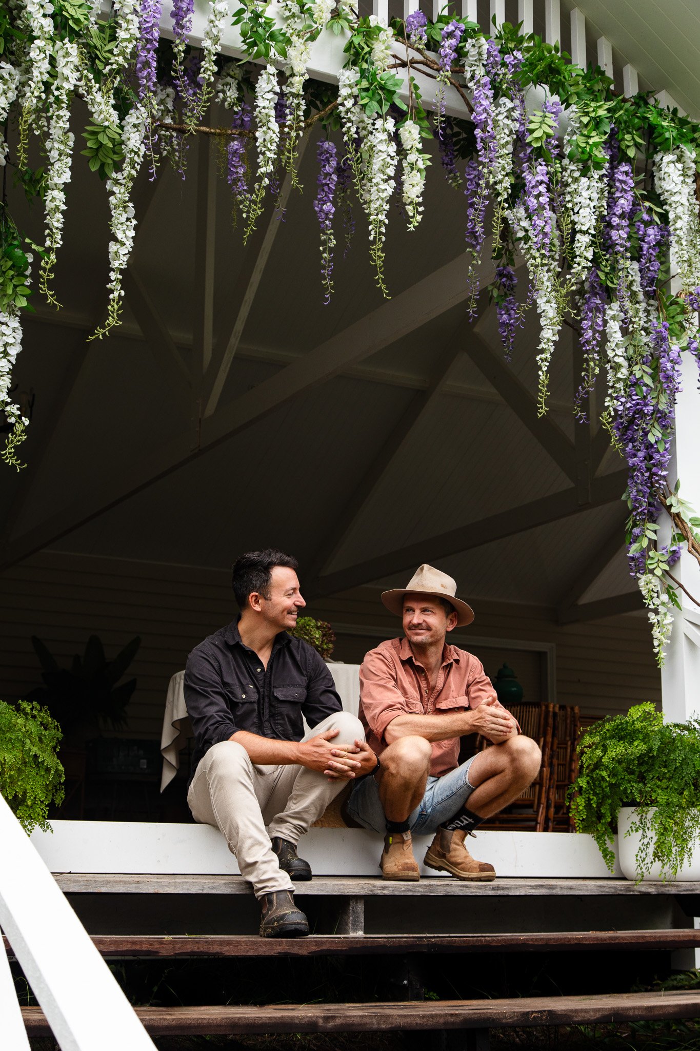 Two men sitting on a porch underneath purple and white hanging flowers and greenery, smiling and engaging in conversation.
