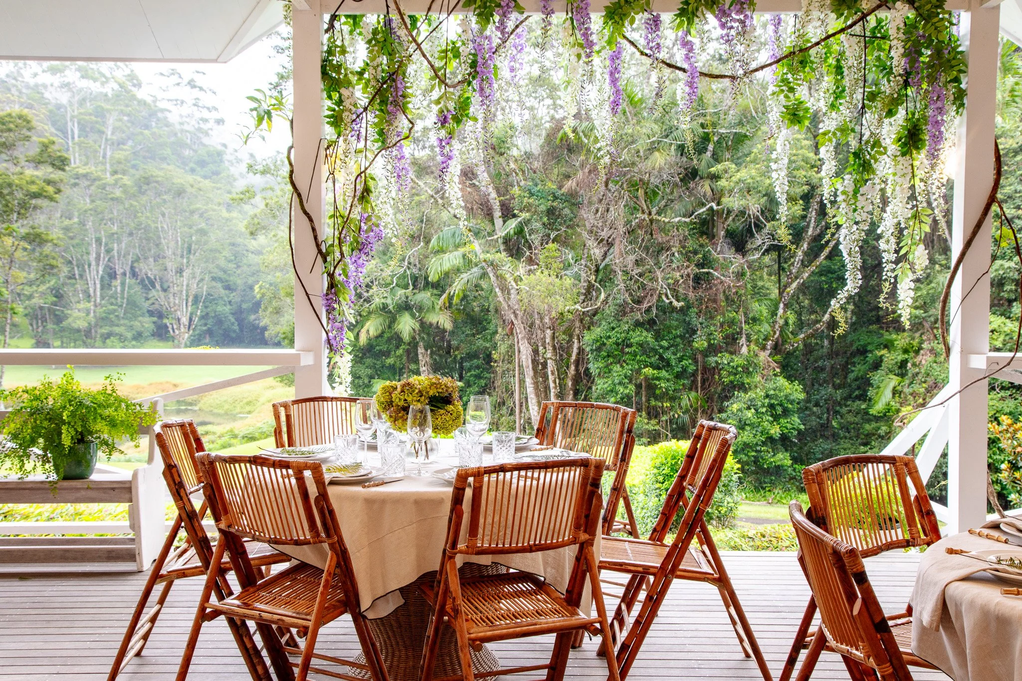Outdoor dining area with a round table, wooden chairs, and a lush, green forest background. The table is set with glasses, plates, and a floral centerpiece, framed by hanging purple and white flowers.