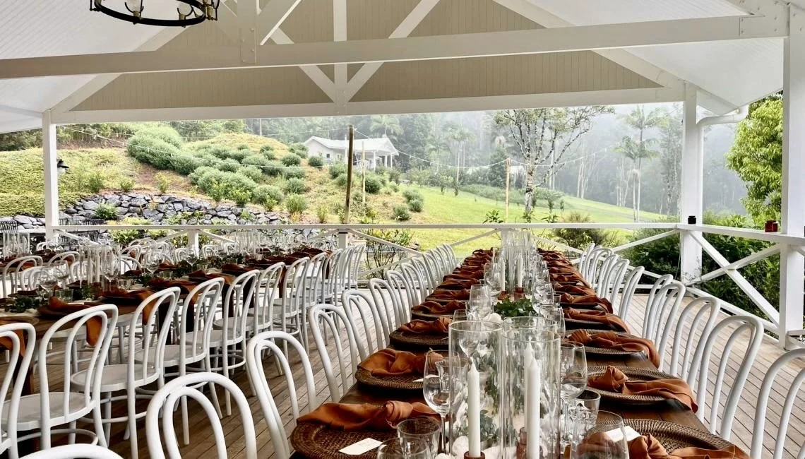 Set dining table with white chairs, glassware, candles, and floral centerpieces under a covered porch overlooking a green hillside with trees and shrubs.