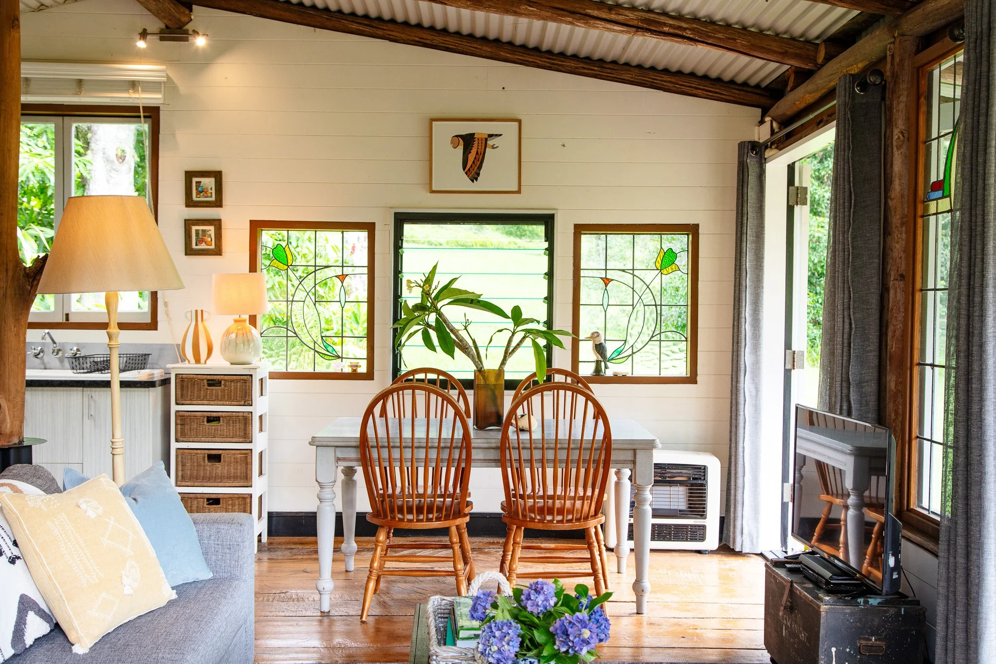 Interior view of a cozy home with a white wooden wall, with a dining table and chairs, a sofa with cushions, a TV on a stand, framed artwork, and windows with green scenery outside.