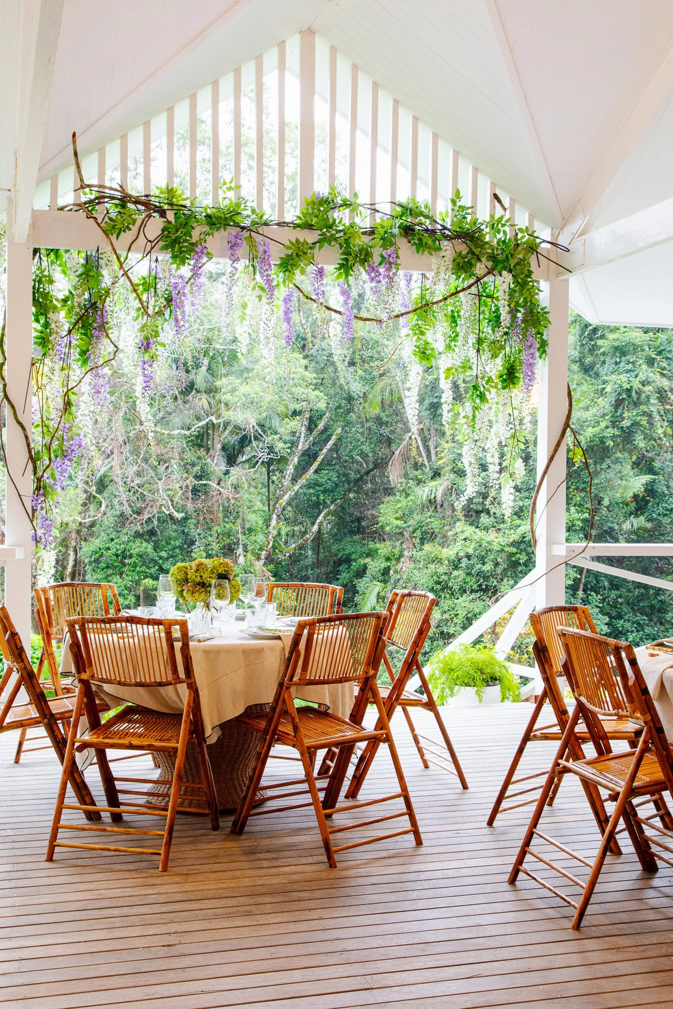 Outdoor dining area with a round table set with a white tablecloth, glassware, and a floral centerpiece, surrounded by wooden chairs, on a wooden deck with lush green trees in the background, and purple and white wisteria hanging overhead.