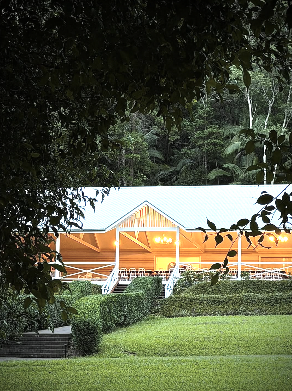 A house with a white roof, beige walls, and a porch with white railings, lit by warm yellow lights, situated in a lush green area with well-kept lawn and bushes, partially obscured by tree branches.