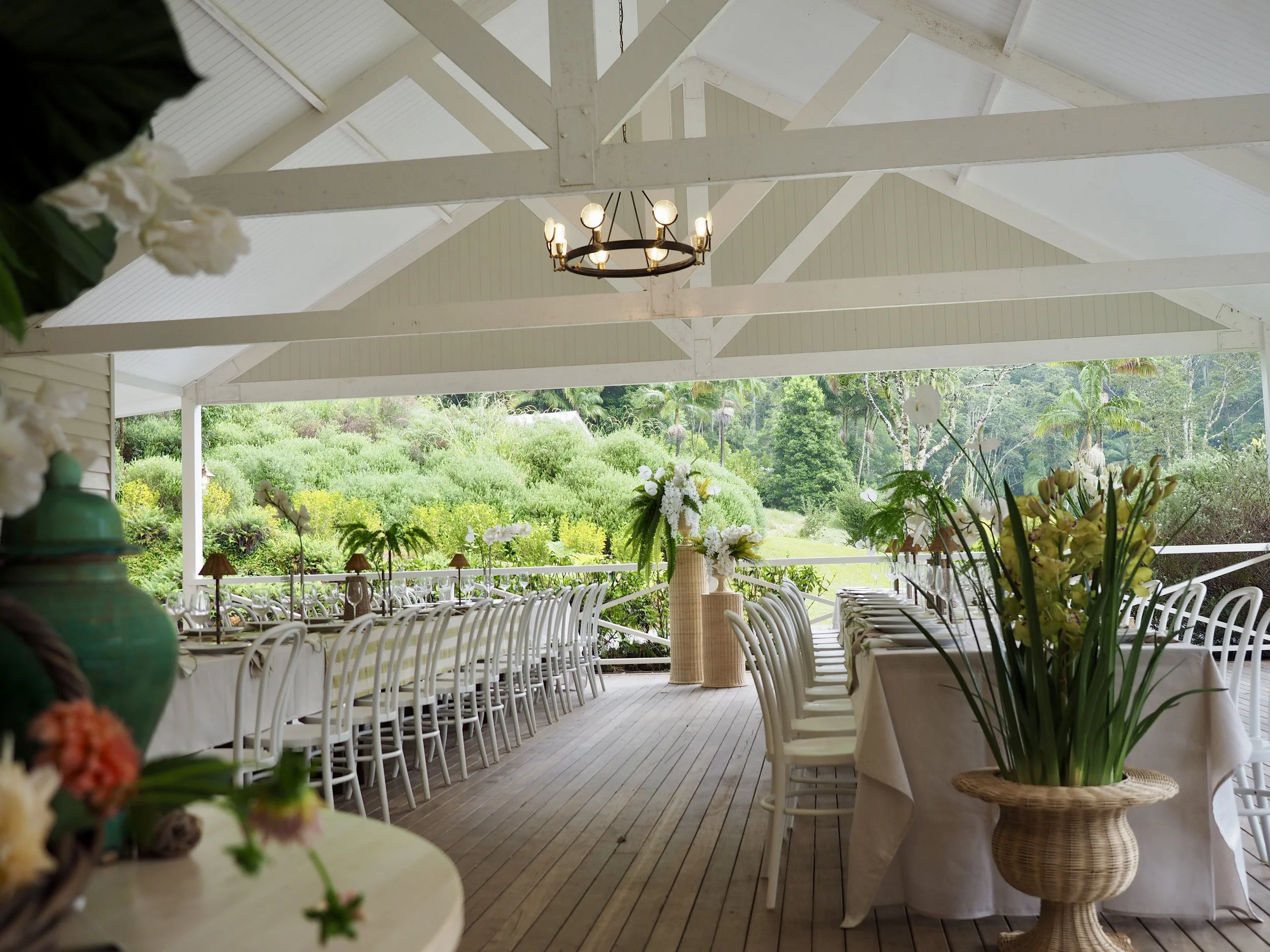 An outdoor wedding or event venue with white tables and chairs, decorated with floral centerpieces and surrounded by lush greenery and trees.