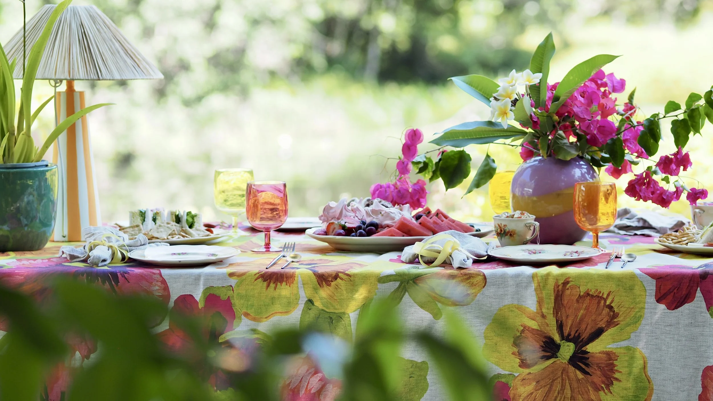 A colorful outdoor dining table set with floral tablecloth, vases of pink and white flowers, glasses, plates, napkins, and a platter of fruit, surrounded by greenery.