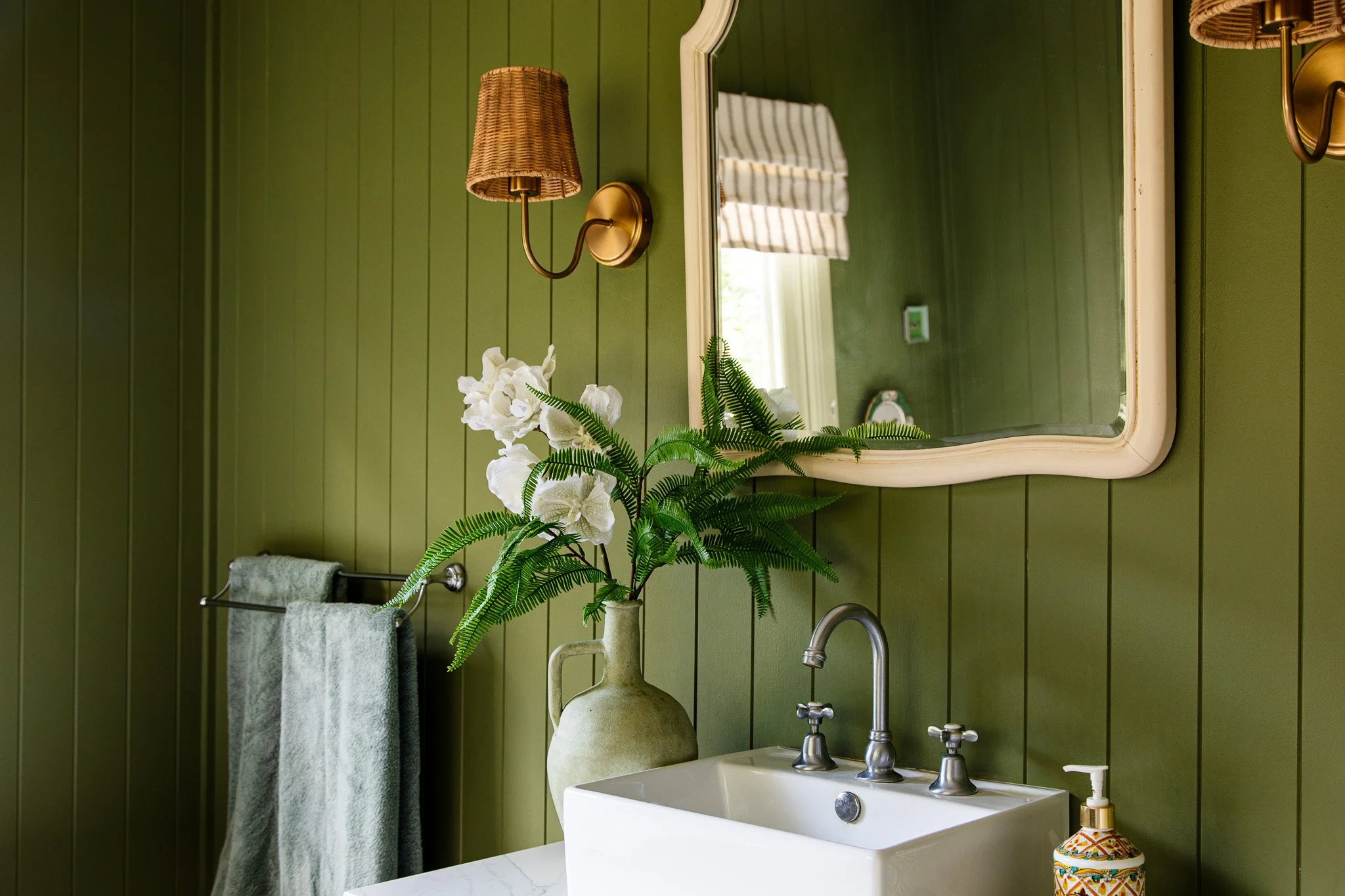 Bathroom with green paneled walls, white sink, mirror, wall lamp, towel rack with gray towels, green vase with white orchids and fern, soap dispenser, and window with striped curtain.