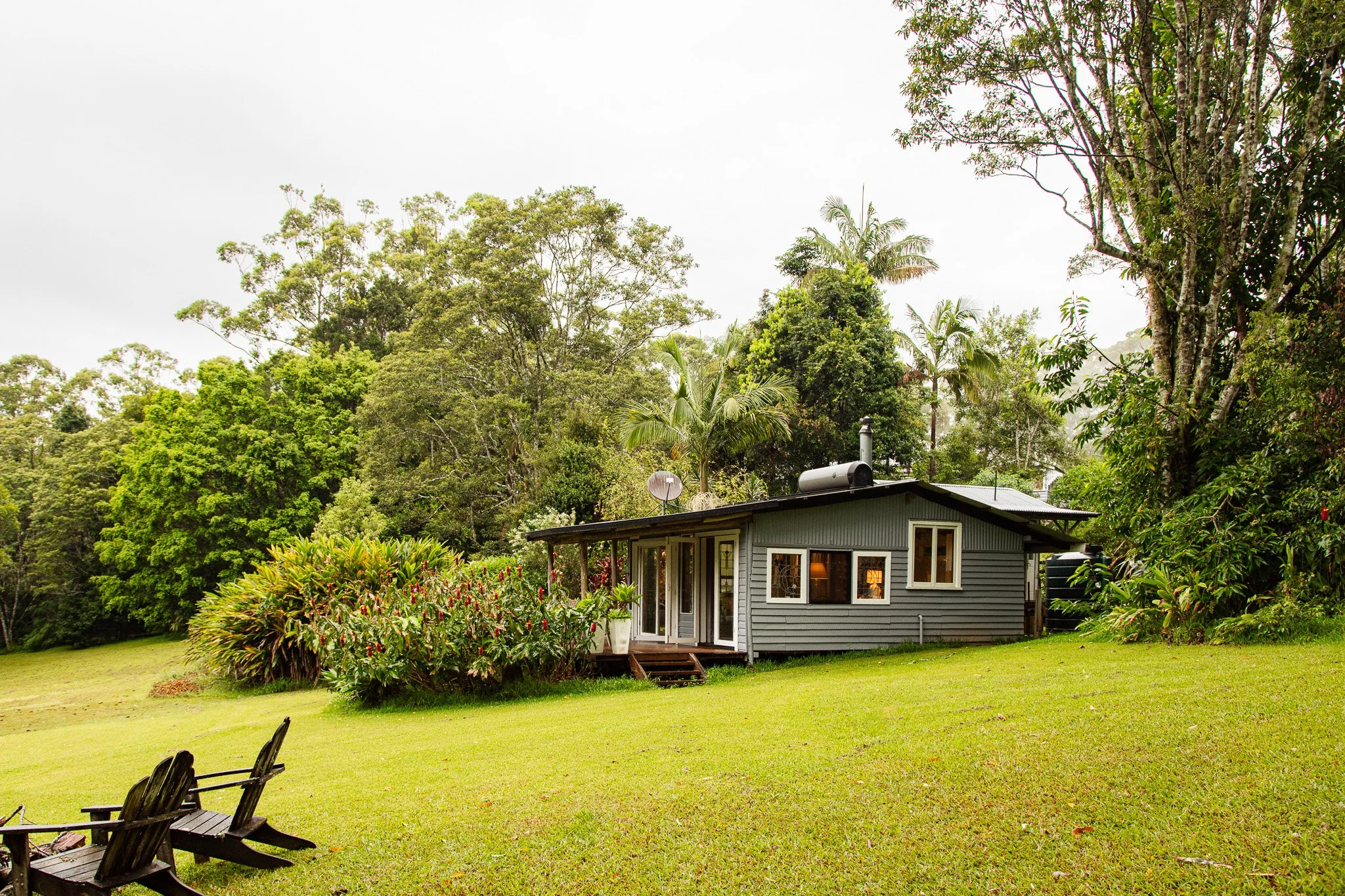 A small gray house with a metal roof sitting in a lush green yard with trees and bushes, two wooden chairs are in the foreground.