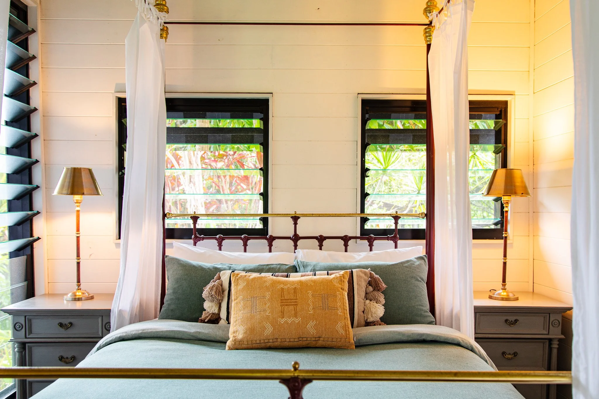 A cozy bedroom with a bed, decorative pillows, and side tables with lamps. Two windows with black frames and bamboo blinds are behind the bed, and white curtains are on either side.