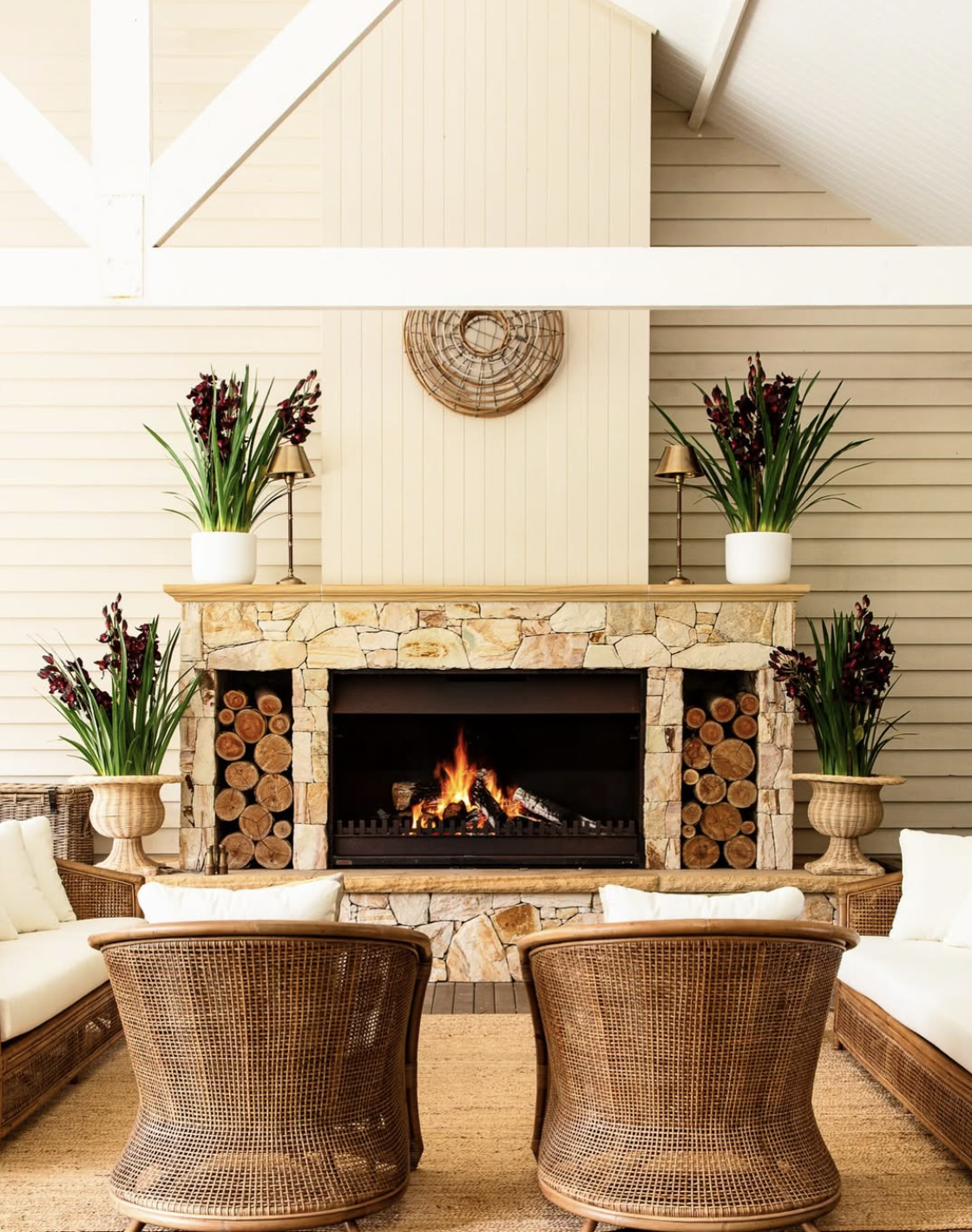 Living room with a stone fireplace, wooden logs, potted plants, and wicker chairs.