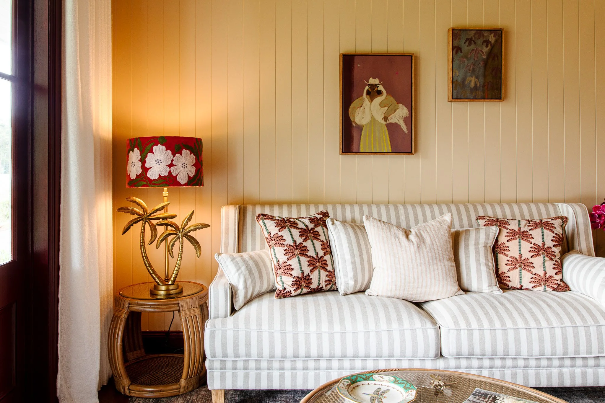 A living room with a striped cream and white sofa decorated with various pillows, a rattan side table with a palm tree lamp, a beige paneled wall with two framed paintings, curtains by a window, and a glass-top coffee table.