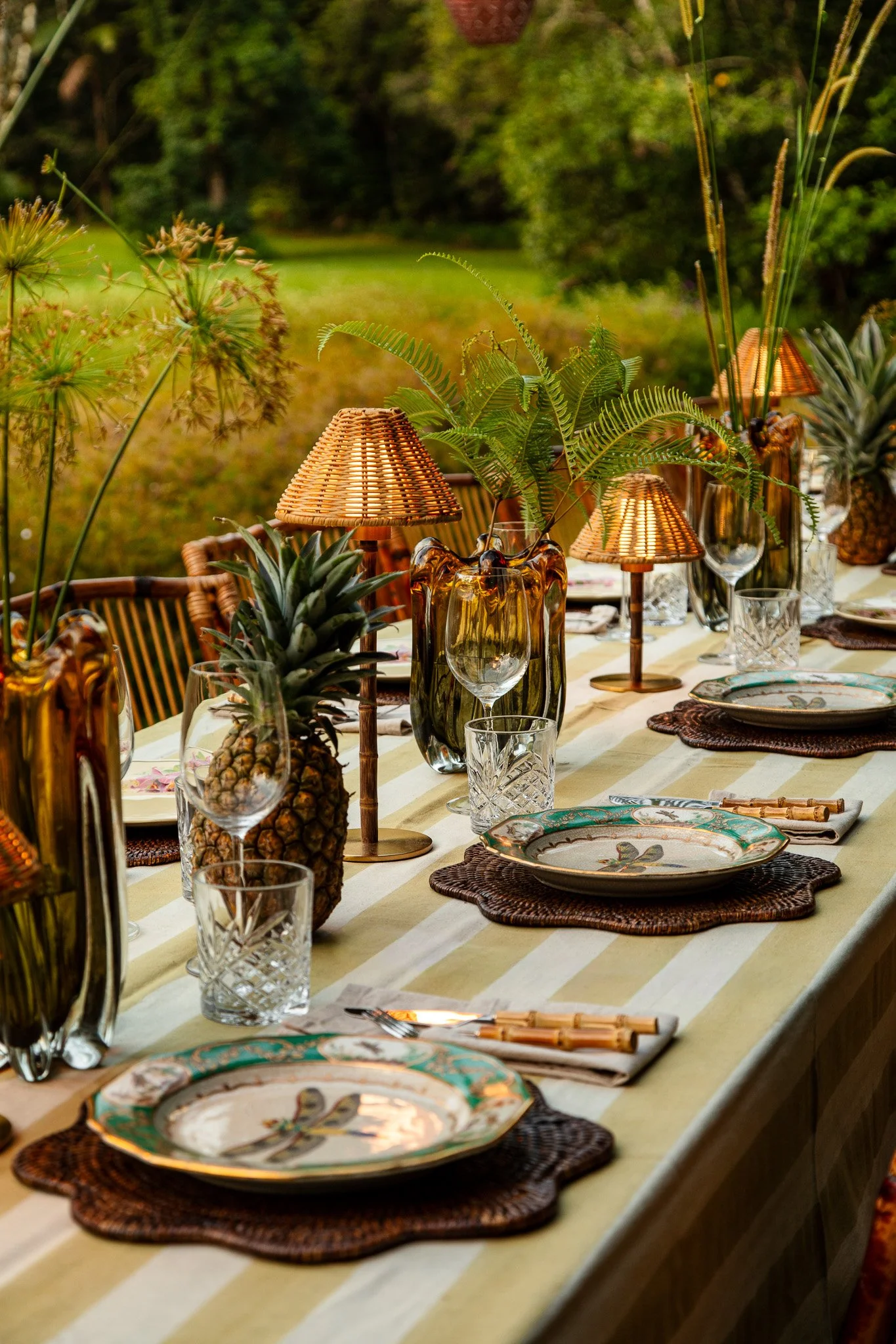 Table setting outdoors with plates, glasses, pineapples, and decorative lamps amidst greenery.