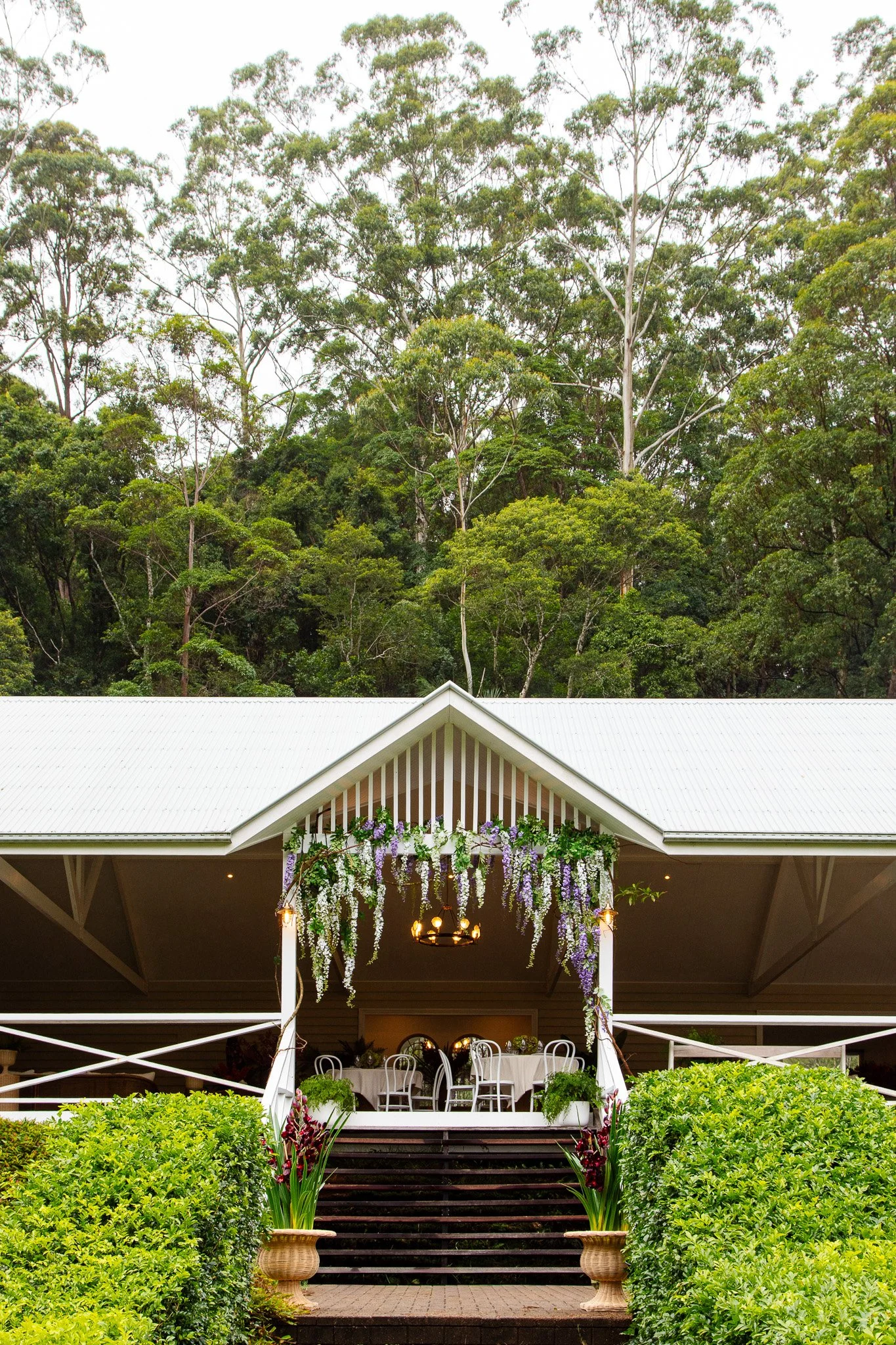 An outdoor reception area with a white porch decorated with hanging purple and white flowers, a chandelier, and a dining setup with white chairs and tables, surrounded by lush green bushes and trees.