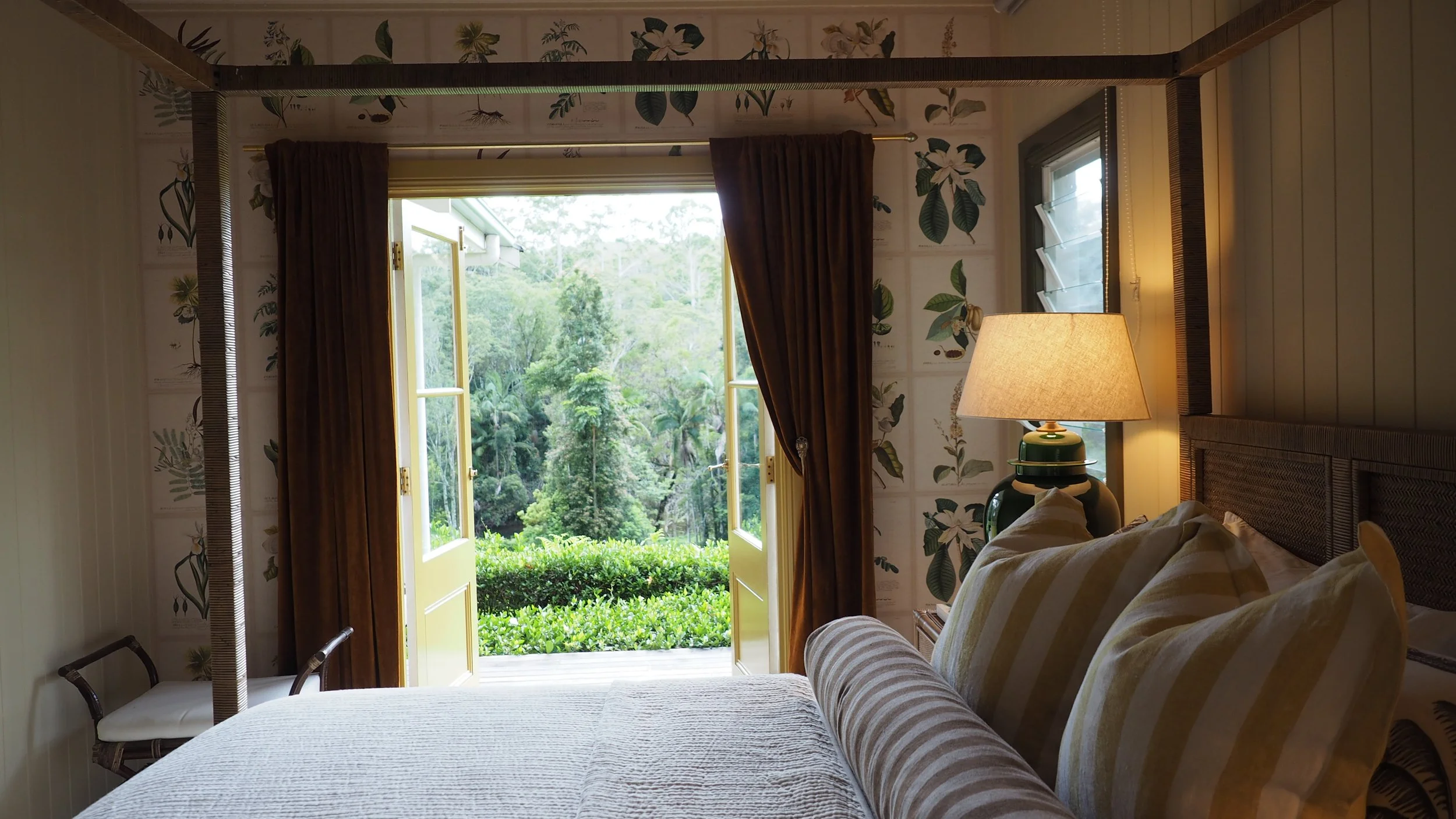 A bedroom with an open door leading to a lush green garden or forest outside, a bed with striped pillows, a lamp on a side table, and floral wallpaper.