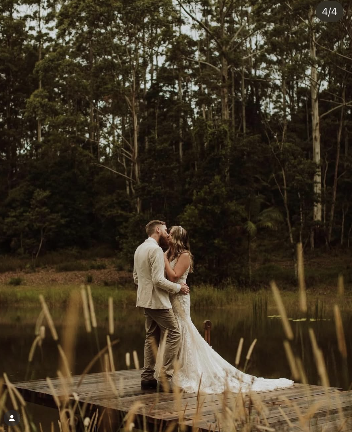 A couple in wedding attire sharing a kiss on a wooden dock at sunset, surrounded by a natural landscape with trees and a pond.