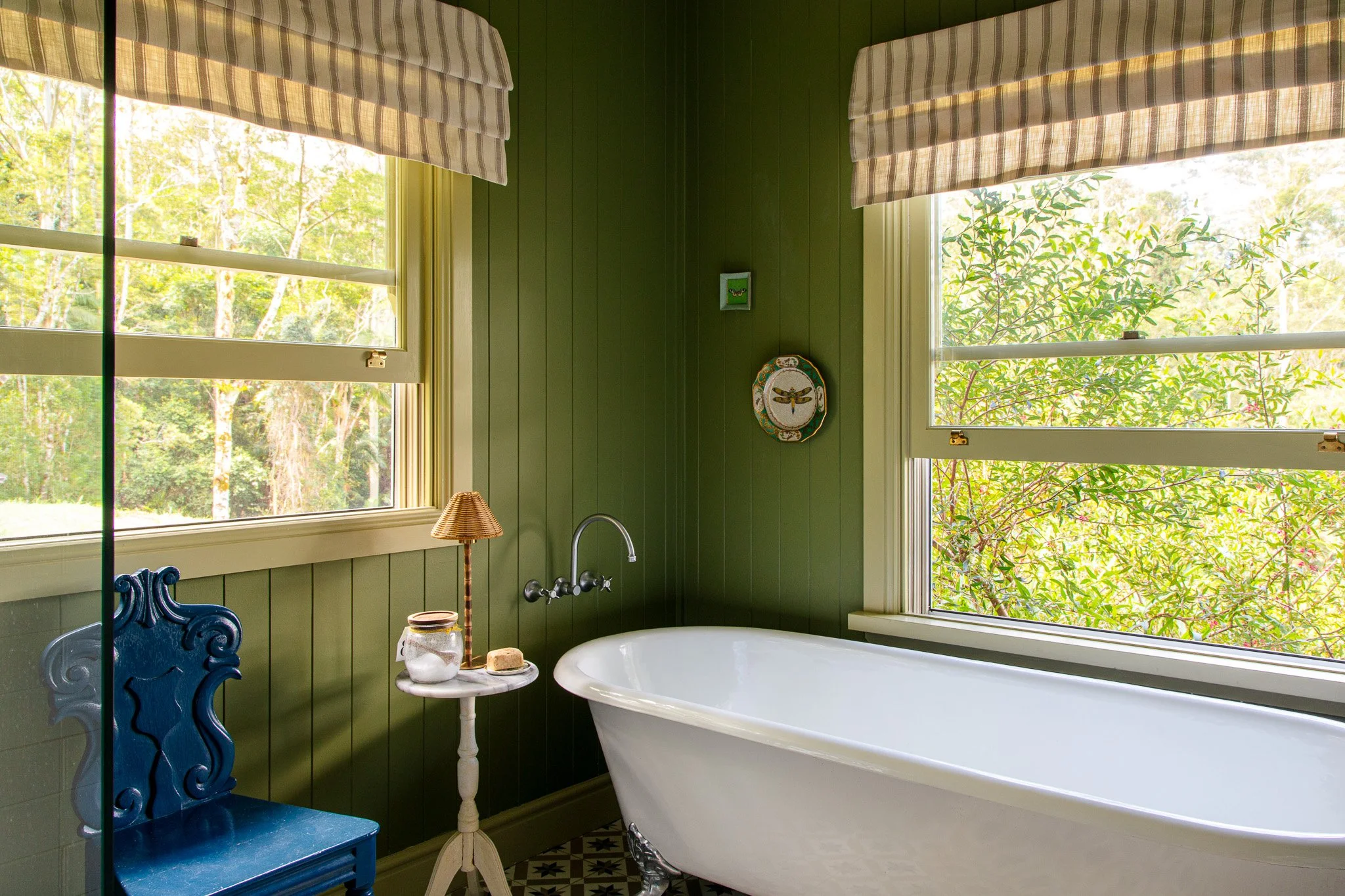 Bathroom with a white clawfoot bathtub, green wooden walls, and two large windows with striped blinds allowing natural light to enter, featuring outdoor greenery.