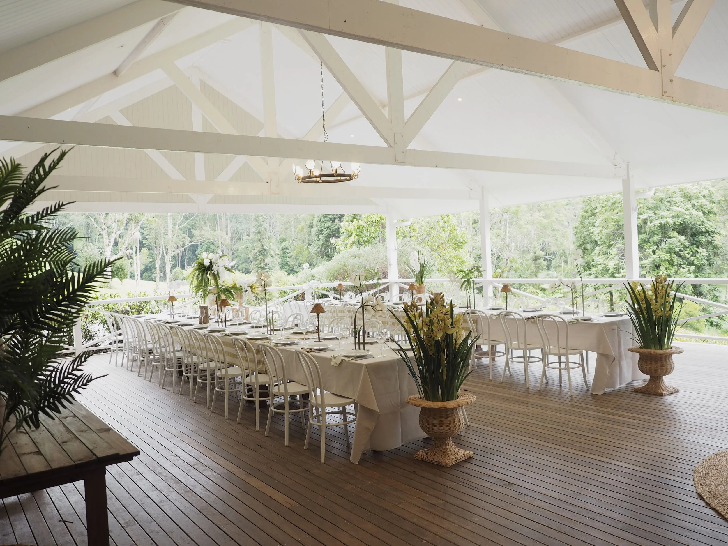 Sunlit outdoor dining area with white tables and chairs, decorated with flowers, under a white pavilion with wooden beams, overlooking lush green trees.
