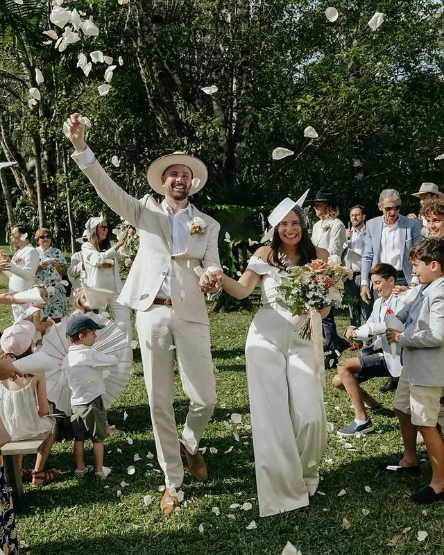 Wedding couple walking hand in hand through guests throwing flower petals, outdoor setting with trees, bride holding bouquet, groom wearing light suit and hat, guests dressed in semi-formal attire celebrating