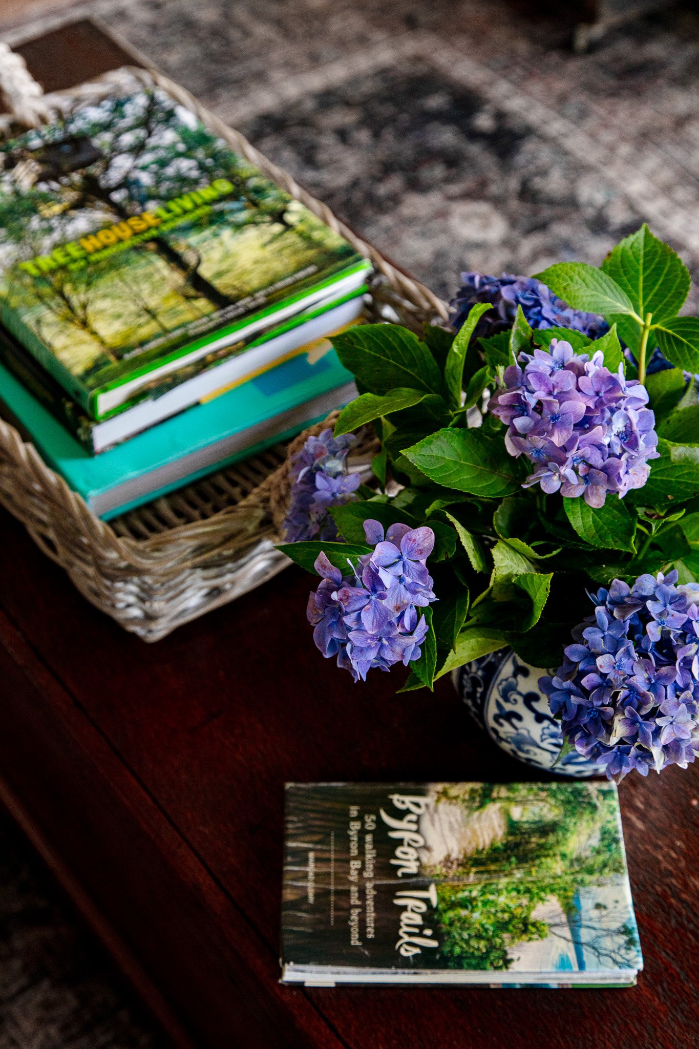 A wicker basket containing gardening books, purple and blue hydrangea flowers in a decorative blue and white vase, and a small booklet titled "Tuesdays with Toads" on a dark wooden surface.