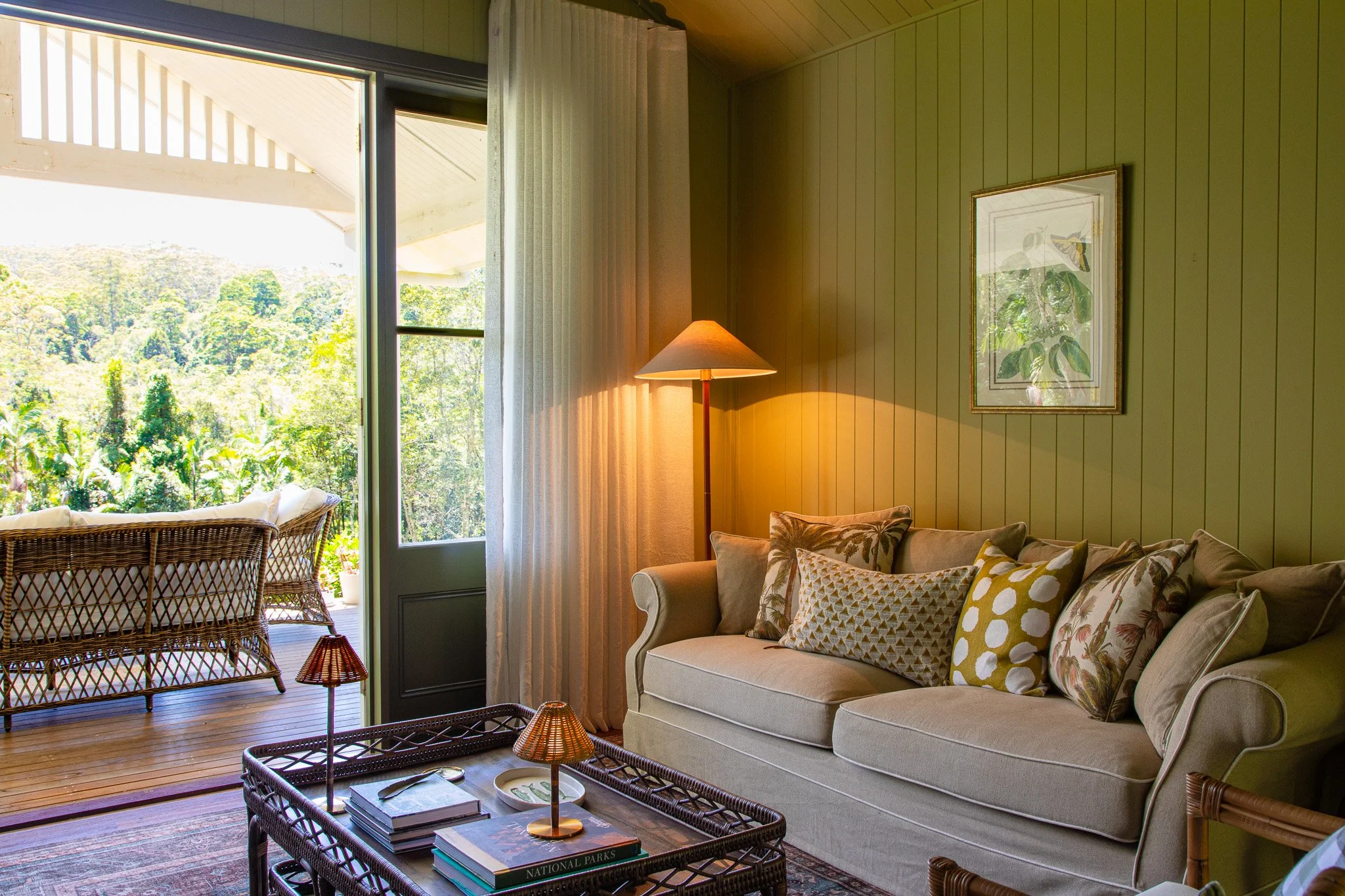 Living room with cream sofa and patterned cushions, floor lamp, framed botanical art, open sliding door leading to a balcony with wicker outdoor furniture and a view of green forest outside.