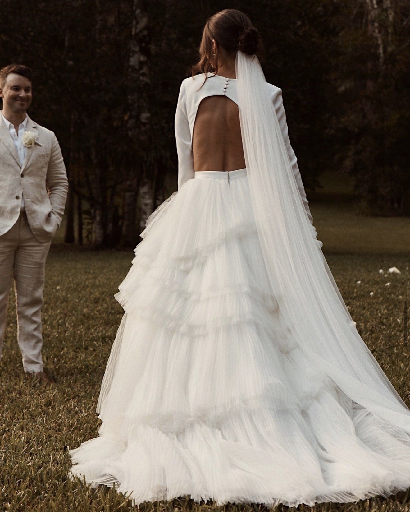 Bride in a white wedding dress with a large ruffled skirt and open back, standing outdoors, with groom in a light-colored suit watching her.