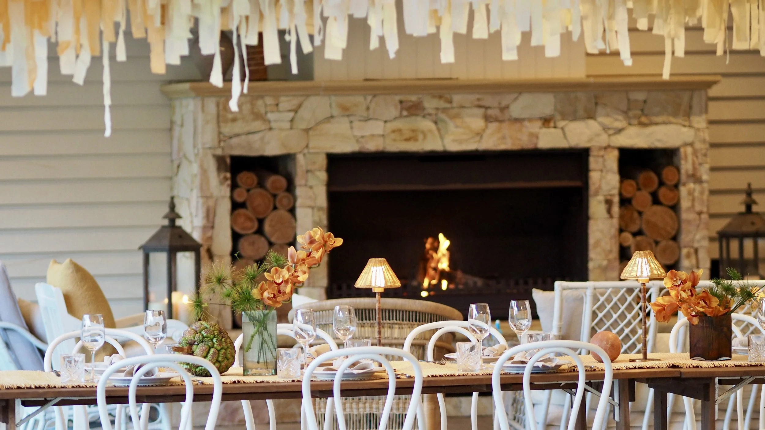 Indoor dining area with a rustic stone fireplace, decorated with vases of orange flowers, table lamps, and set with wine glasses and plates.