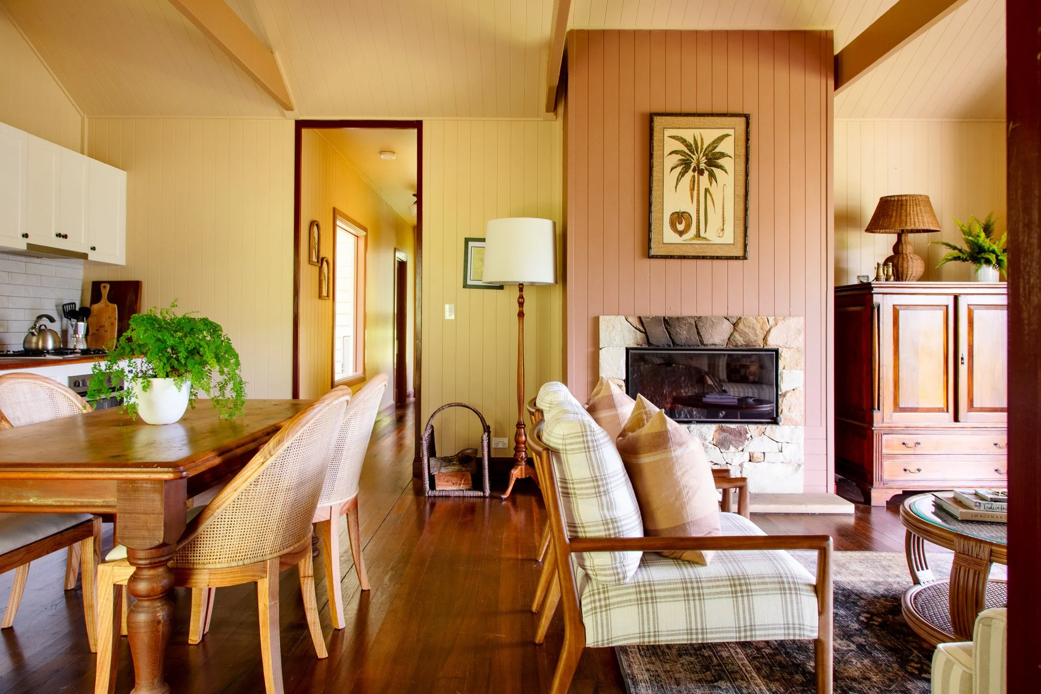 Living room with wood-paneled walls, a fireplace with stone surround, plaid armchair, wooden cabinet with lamp and plant, dining table with chairs and a green potted plant.