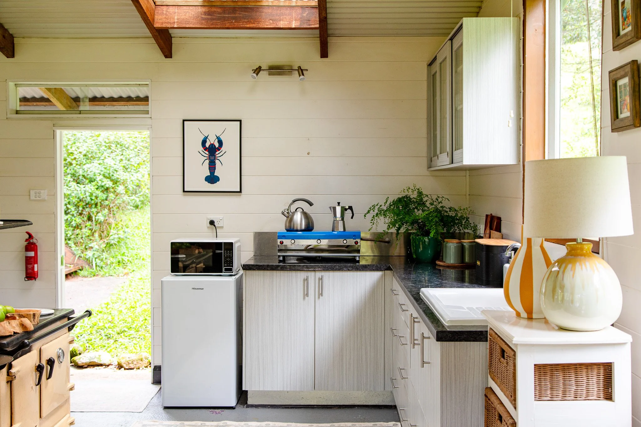 Kitchen with white cabinets, black countertop, and various appliances including a microwave, toaster, and stovetop, with a view of greenery outside through the open door.