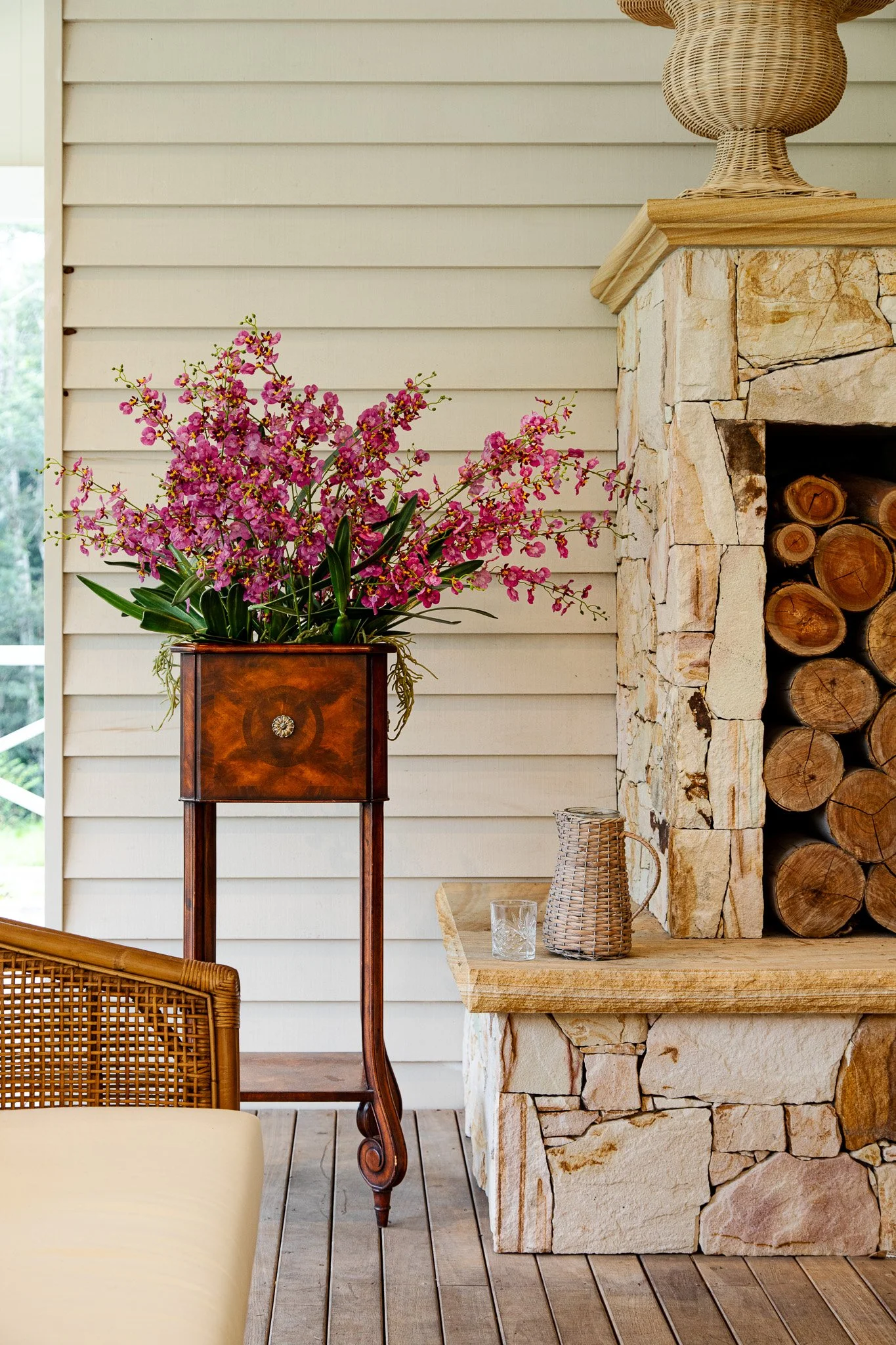 Pink orchid flowers in a wooden plant stand next to a stone fireplace with stacked firewood, with a glass and a woven pitcher on the mantel.