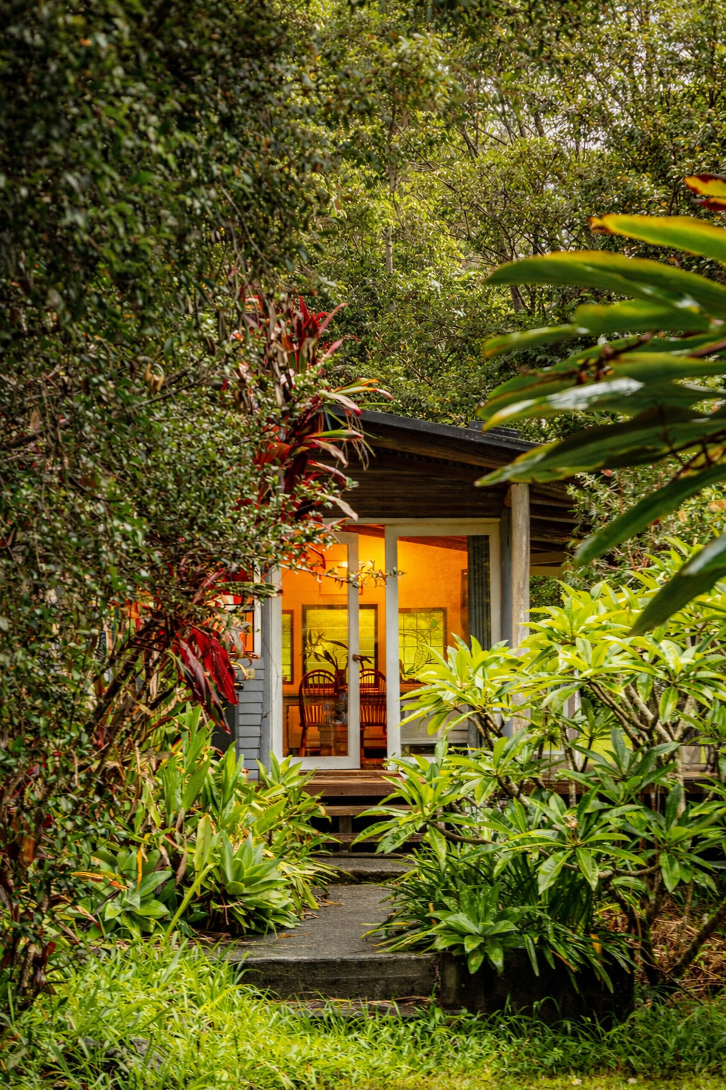 A cozy house with a porch surrounded by lush green plants and trees, with warm yellow light emanating from inside.