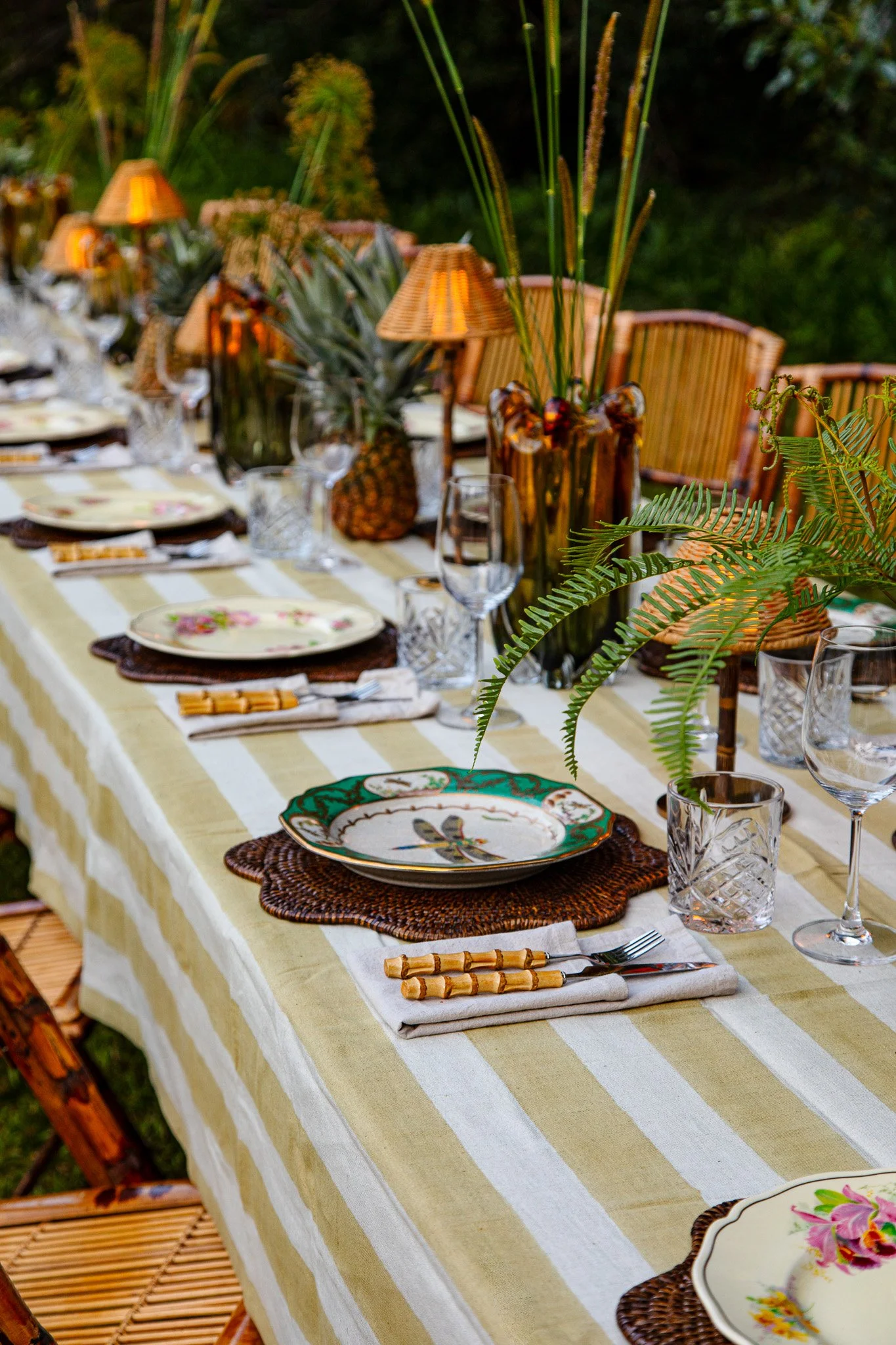 An outdoor dining table set with decorative plates, glassware, cutlery, and woven placemats. The table is adorned with tropical-themed decorations, including pineapples, large vases with green foliage, and small lamps, creating a tropical or garden party ambiance.