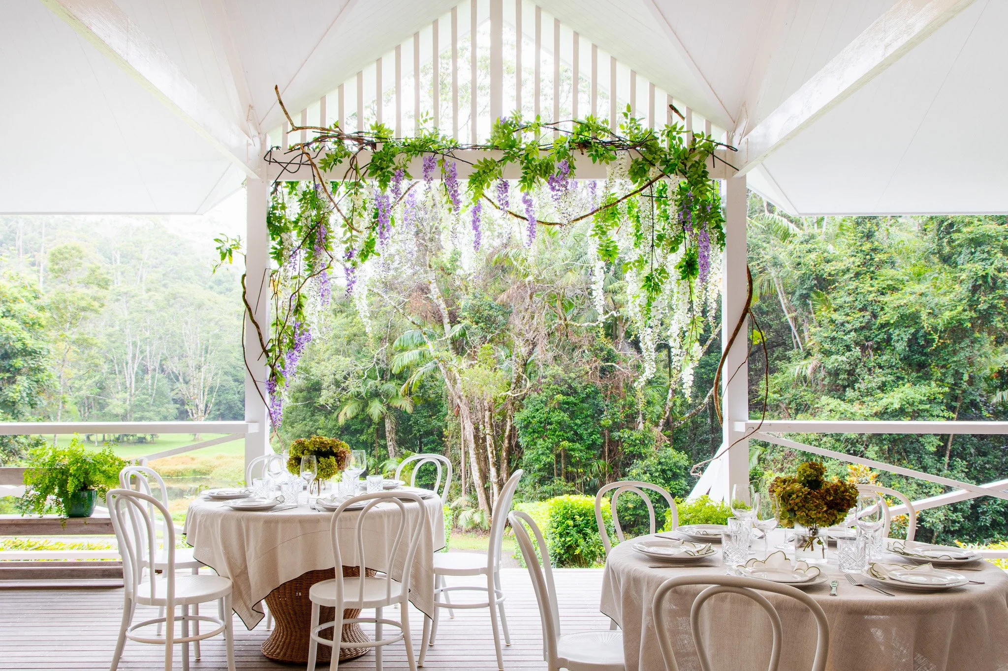 Outdoor dining area with round tables covered in white tablecloths, set with plates, glasses, and floral centerpieces, surrounded by white chairs, with a lush green forest view and hanging purple and white flowers overhead.