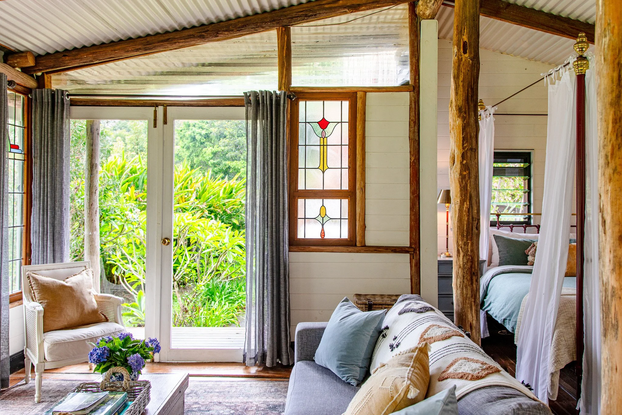Cozy rustic living room with white walls, wooden logs, and a glass door opening to green foliage outside. There is a loveseat with pillows, a side table with books, and a small chair with a cushion. Part of a bedroom with a four-poster bed and white 