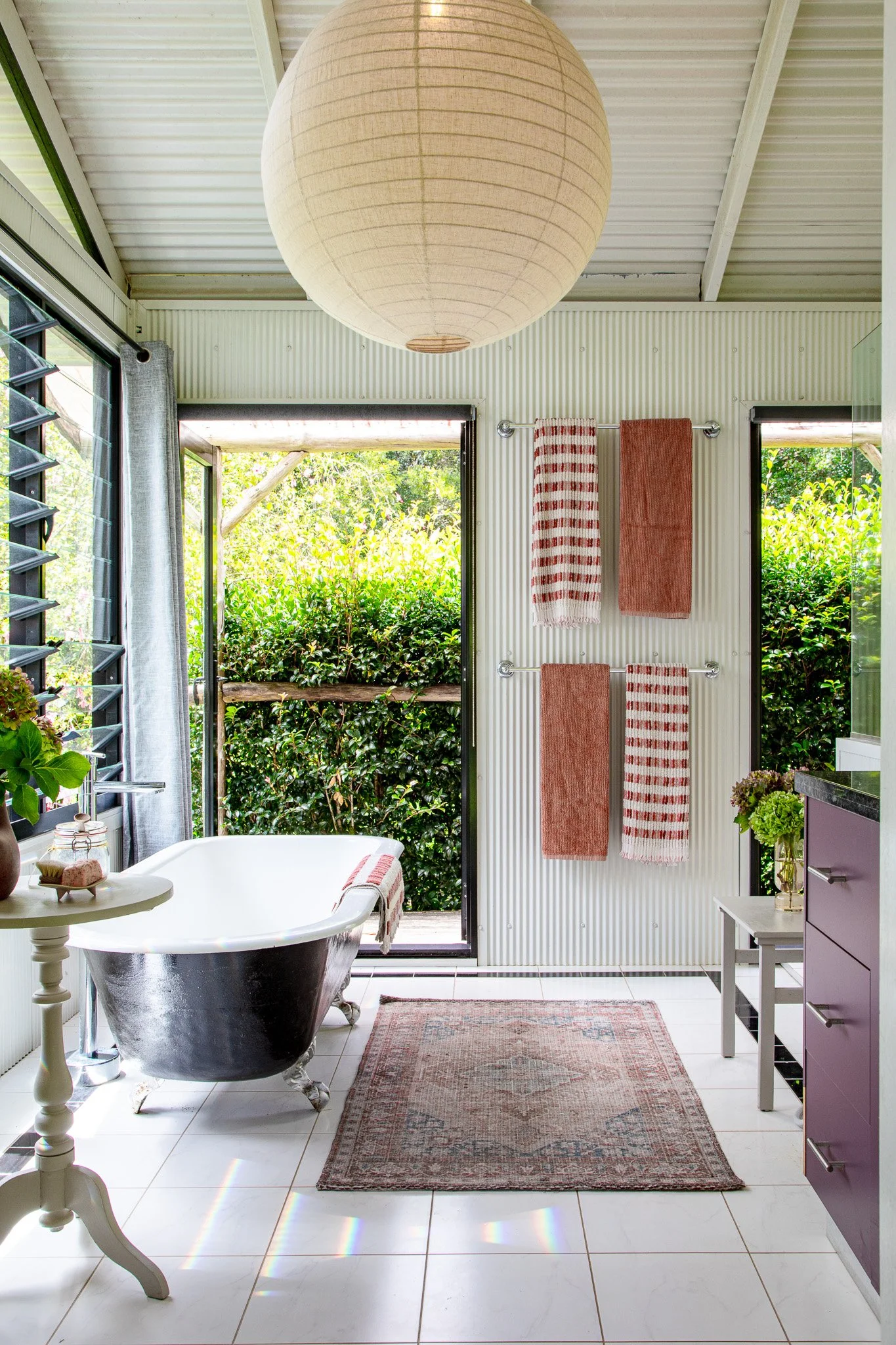 Bright bathroom with corrugated white walls, a clawfoot bathtub, and an open door to a lush green outdoor space. Towel racks with striped and solid towels, a small table with flowers, a purple cabinet, and a vintage rug.