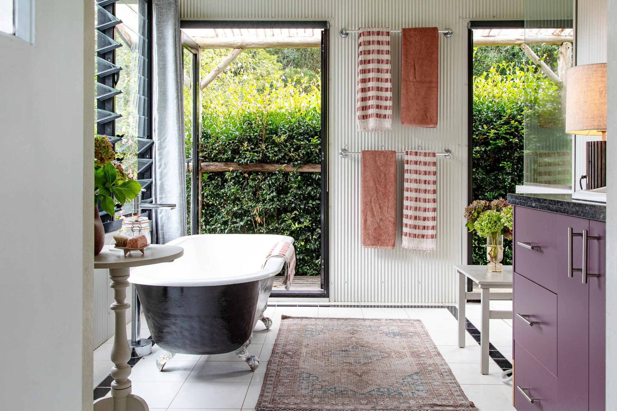 Bathroom with clawfoot bathtub near open door to green outdoor space, pink towels on wall, purple vanity, floral arrangements, patterned rug on white tiled floor.