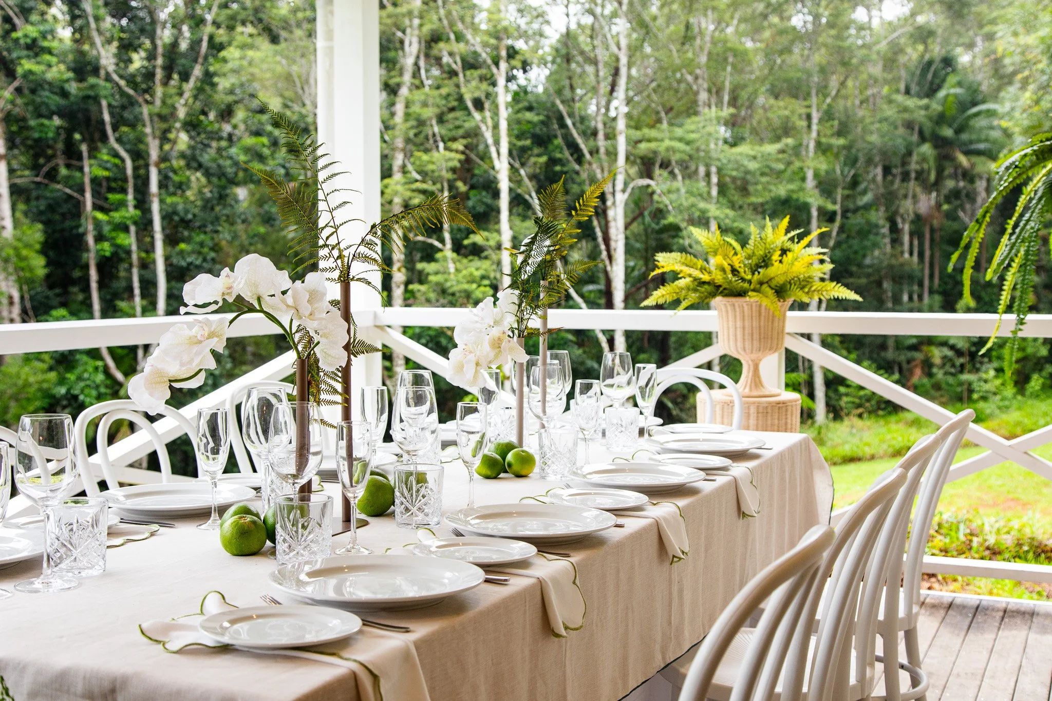 A beautifully set outdoor dining table with white plates, wine glasses, and napkins, decorated with white flowers and greenery, with a lush green forest background.