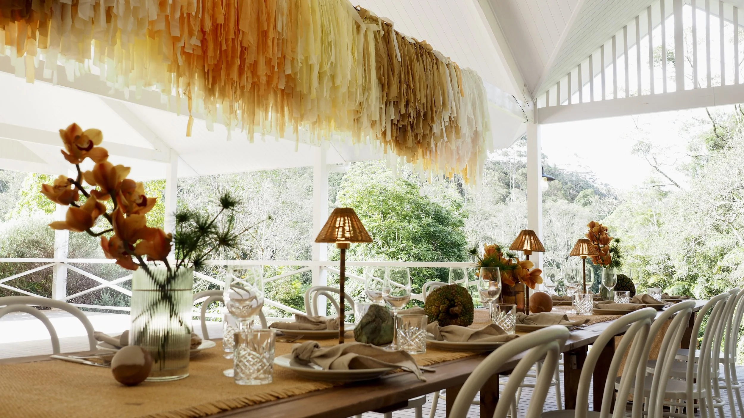 Decorated outdoor dining table with flowers, glasses, and tableware under a canopy with hanging paper decorations and surrounded by lush greenery.