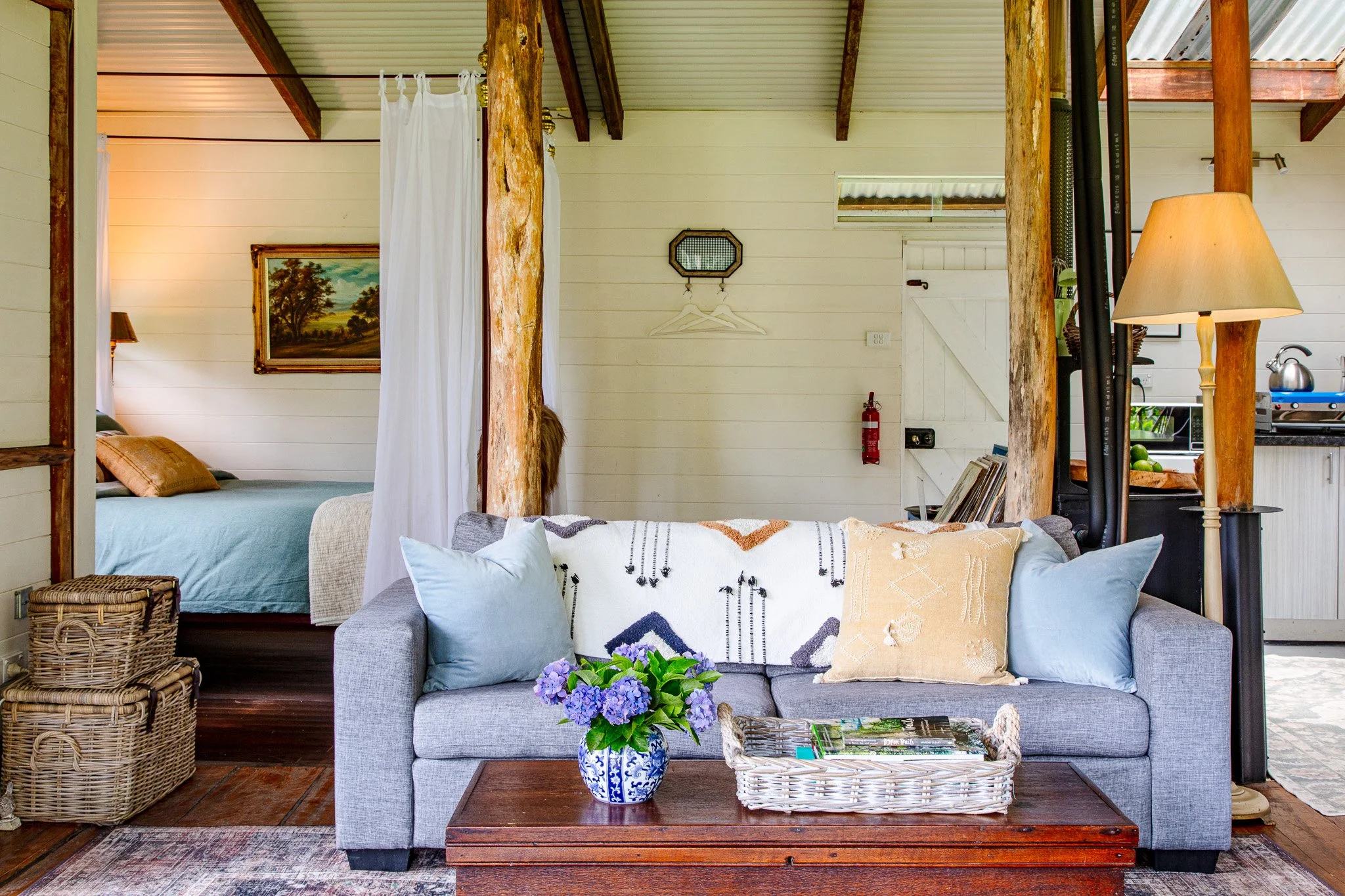 A cozy living room with a gray sofa adorned with light blue and beige pillows, a wooden coffee table with a floral vase and magazines, a white throw blanket, and a rustic interior with wooden beams, white paneled walls, and a view into a bedroom and 