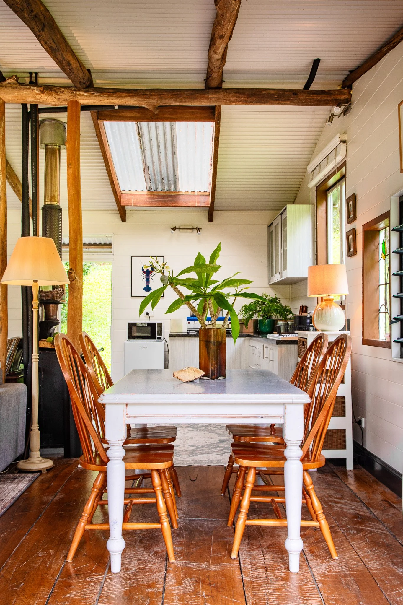 A cozy dining area with a white wooden table, surrounded by wooden chairs, with a potted green plant as a centerpiece. In the background, a kitchen with cabinetry, a microwave, and various decor items is visible. The room has wooden floors and a ceil