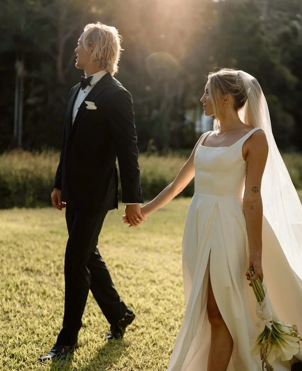 A bride and groom holding hands outdoors during sunset, with the groom in a black tuxedo and the bride in a white wedding dress with a veil, walking on grassy ground.
