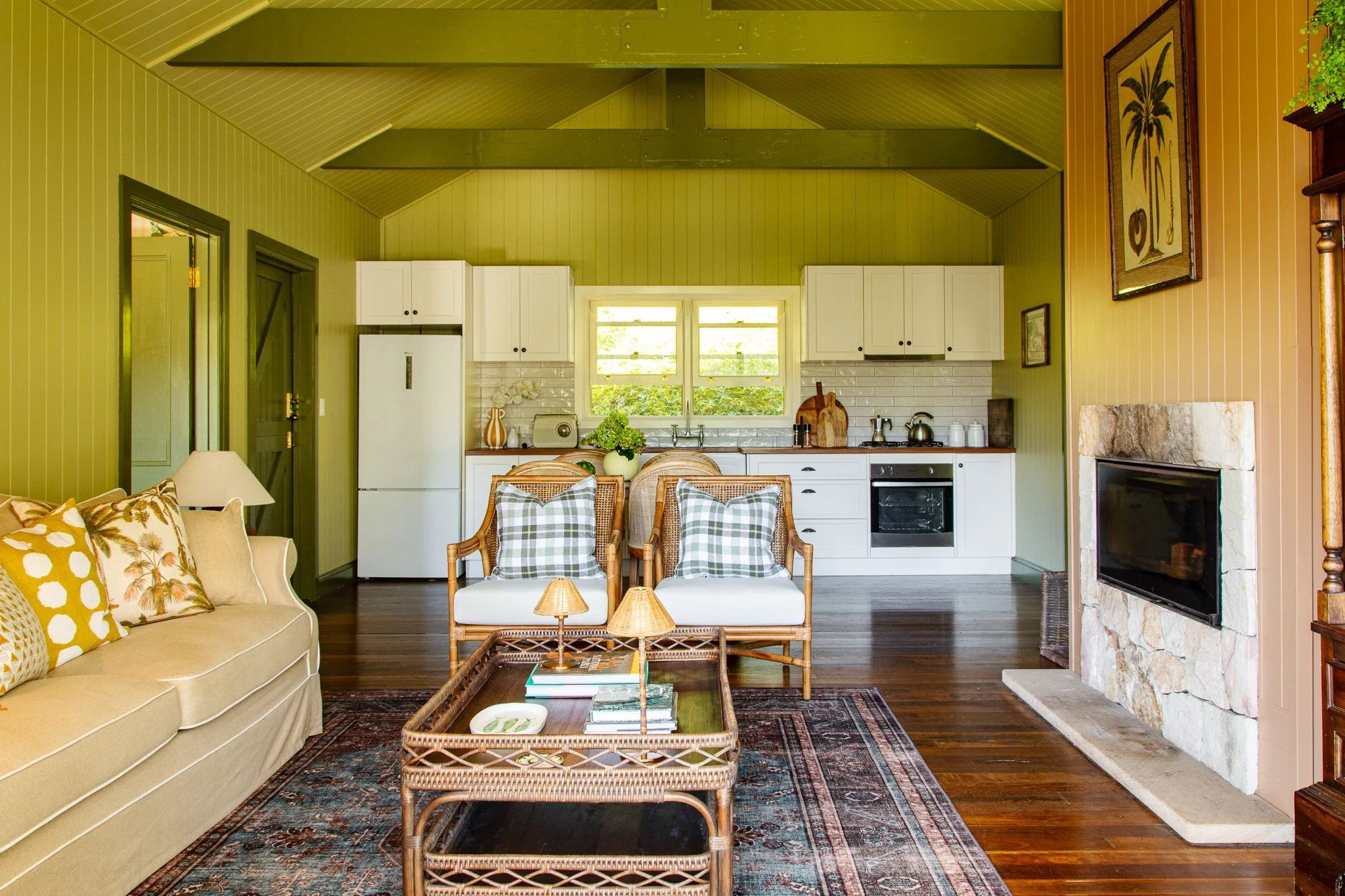 Living room with green walls, wooden floors, fireplace, beige sofa with patterned cushions, and wicker chairs with checkered cushions. Open kitchen in background with white cabinets, window, and kitchen appliances.
